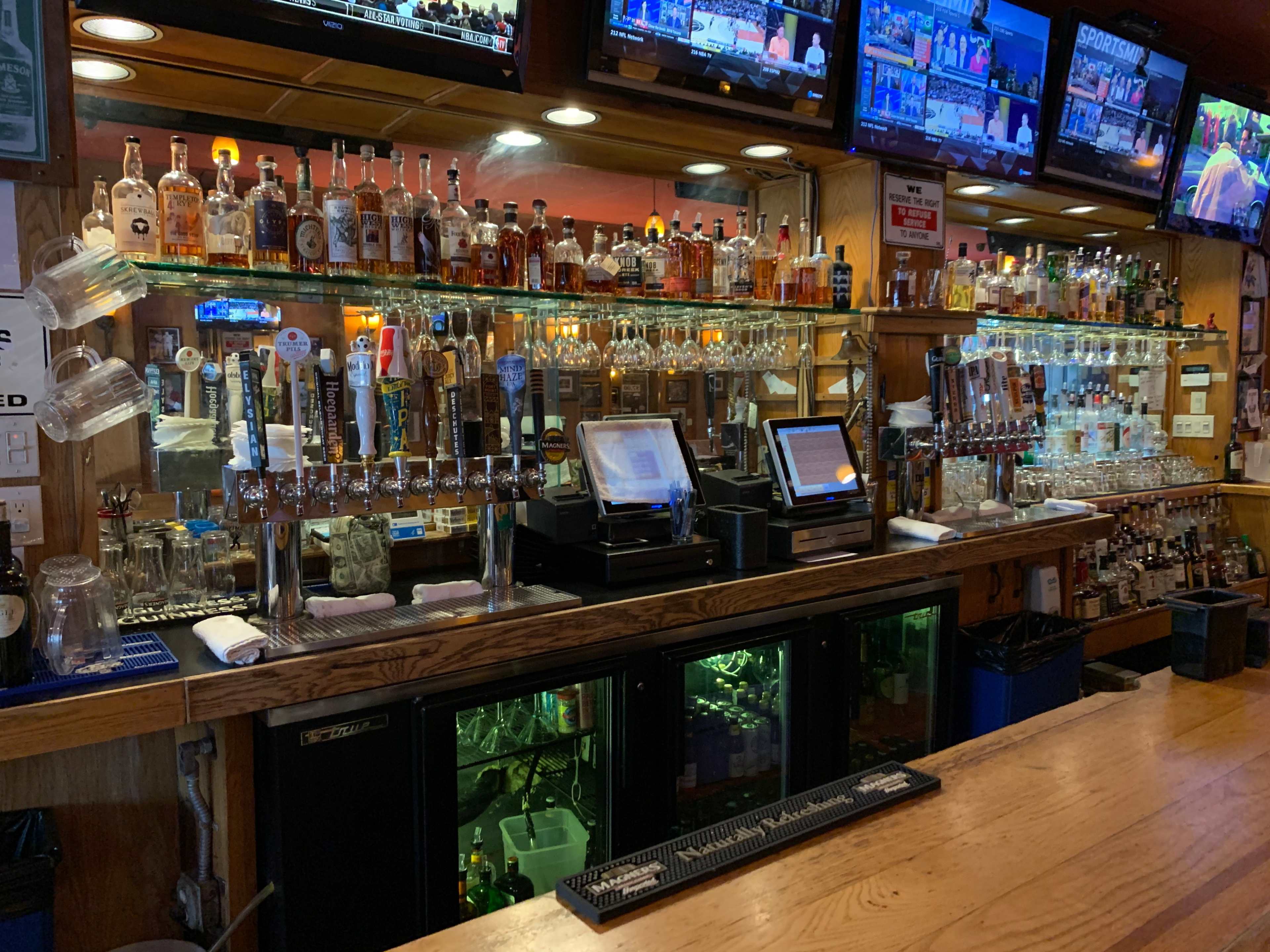 The image shows a well-stocked bar with various bottles of liquor, beer taps, and several television screens displaying sports.