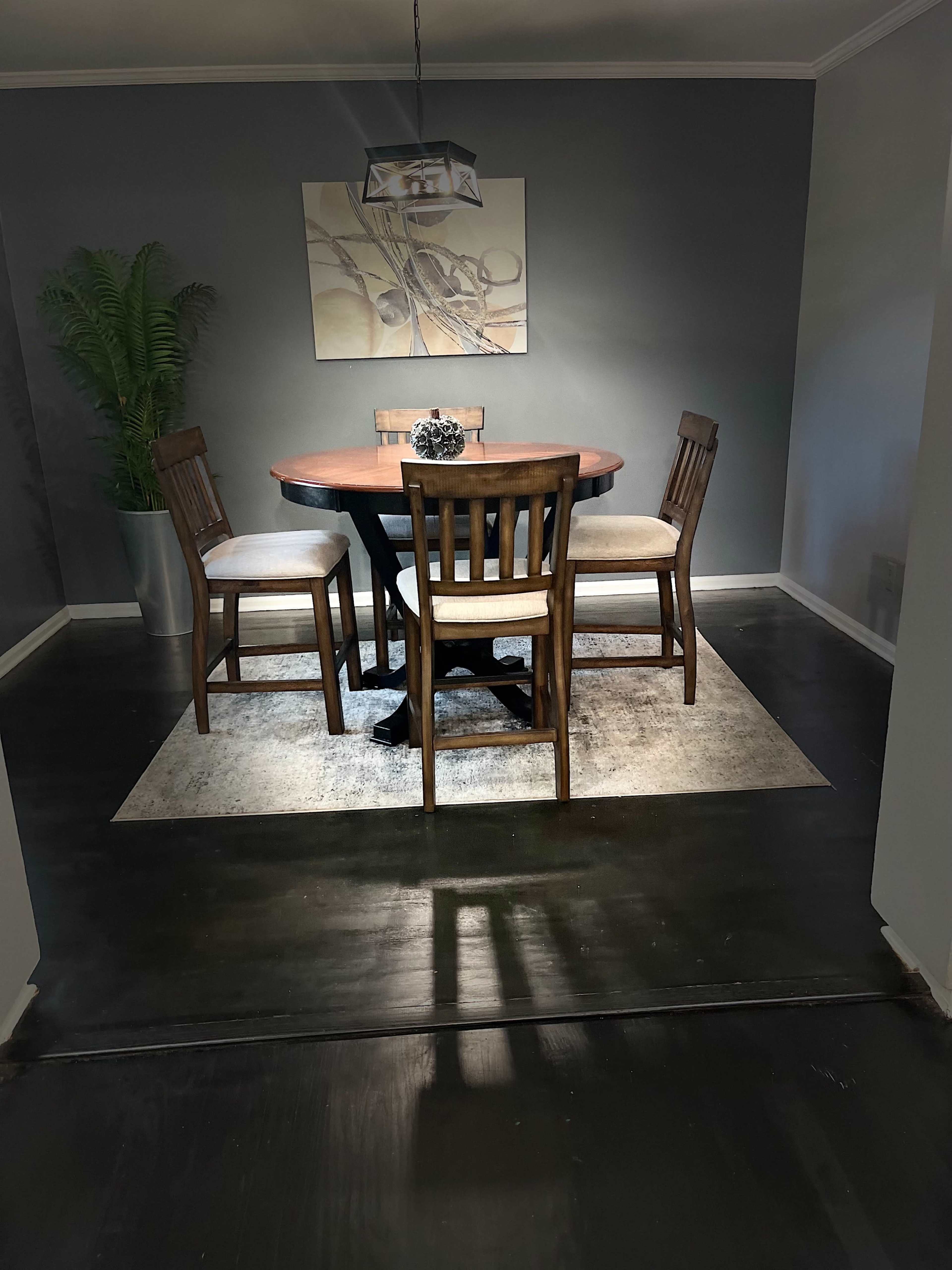 A circular wooden dining table surrounded by four wooden chairs is positioned on a rug in a dimly lit room with gray walls and a potted plant in the corner.