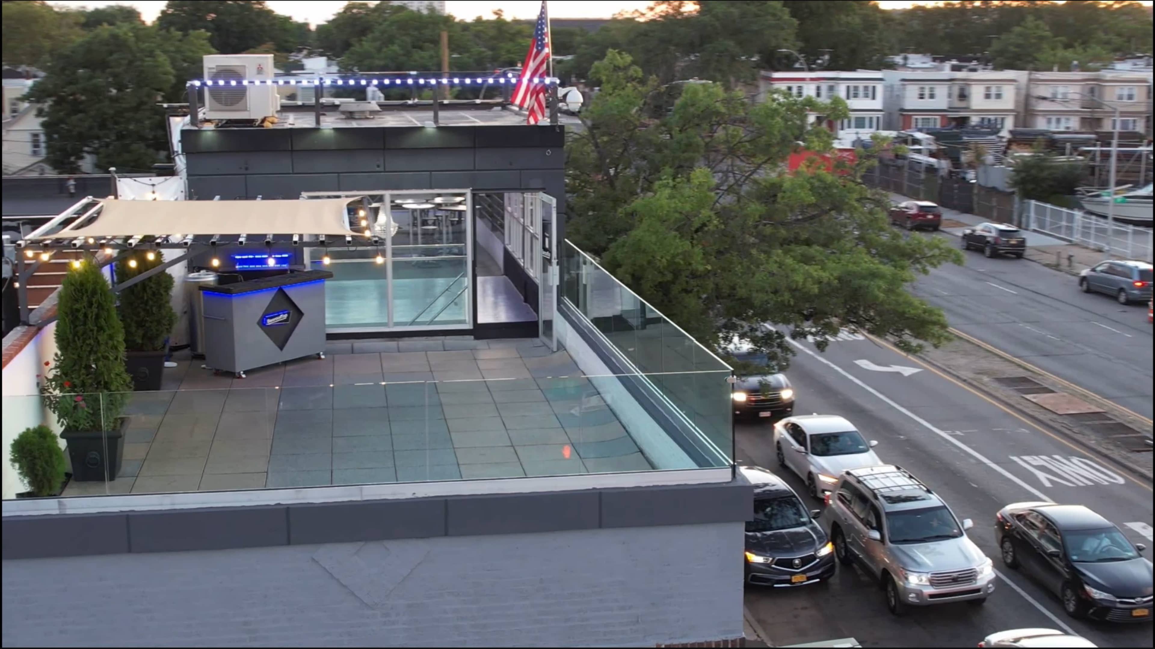 The image shows a rooftop deck with a glass railing, an American flag, and a modern structure overlooking a street with parked cars.