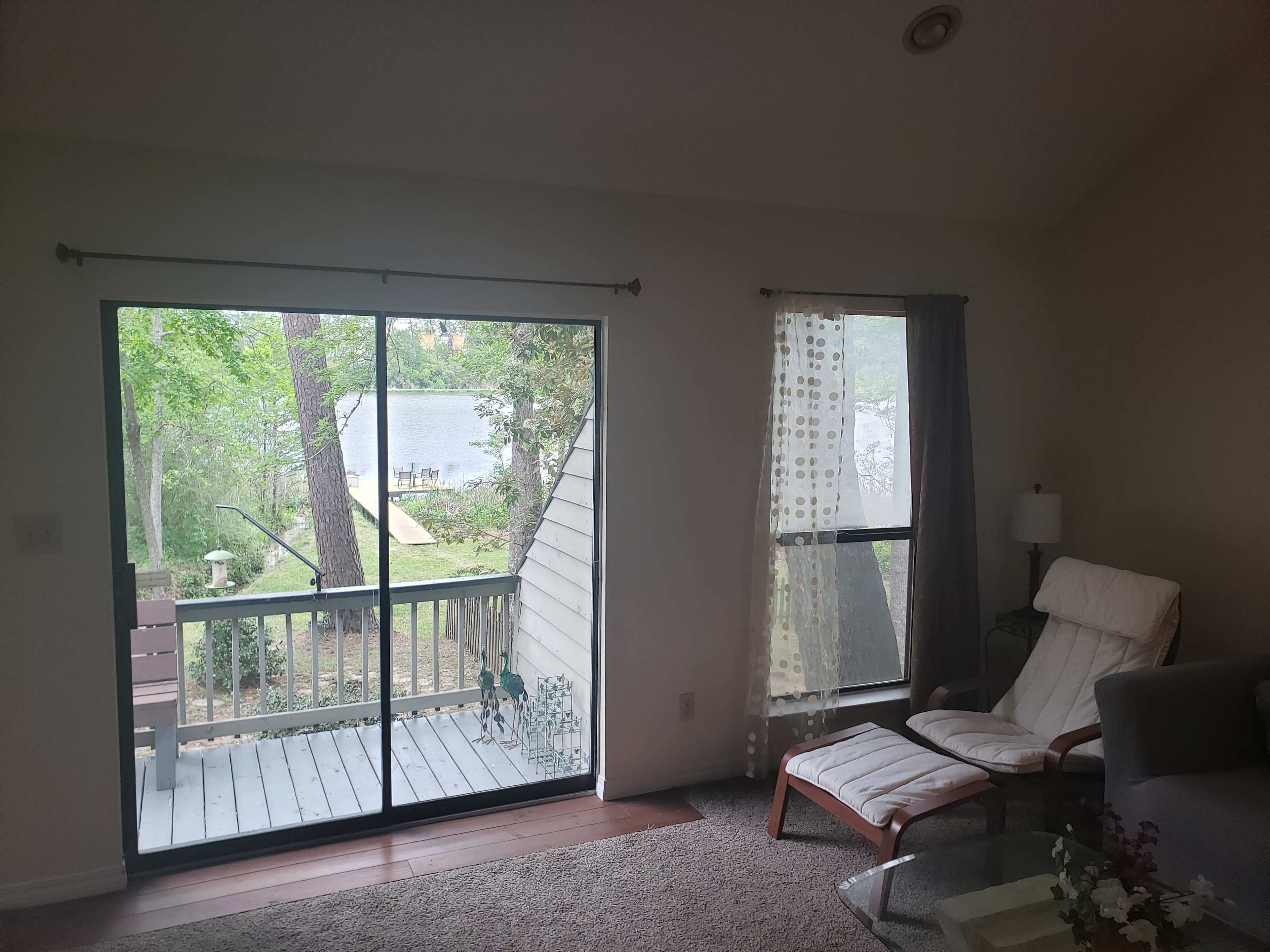 A living room features a large sliding glass door leading to a deck overlooking a lake, with a comfortable chair and lamp nearby.