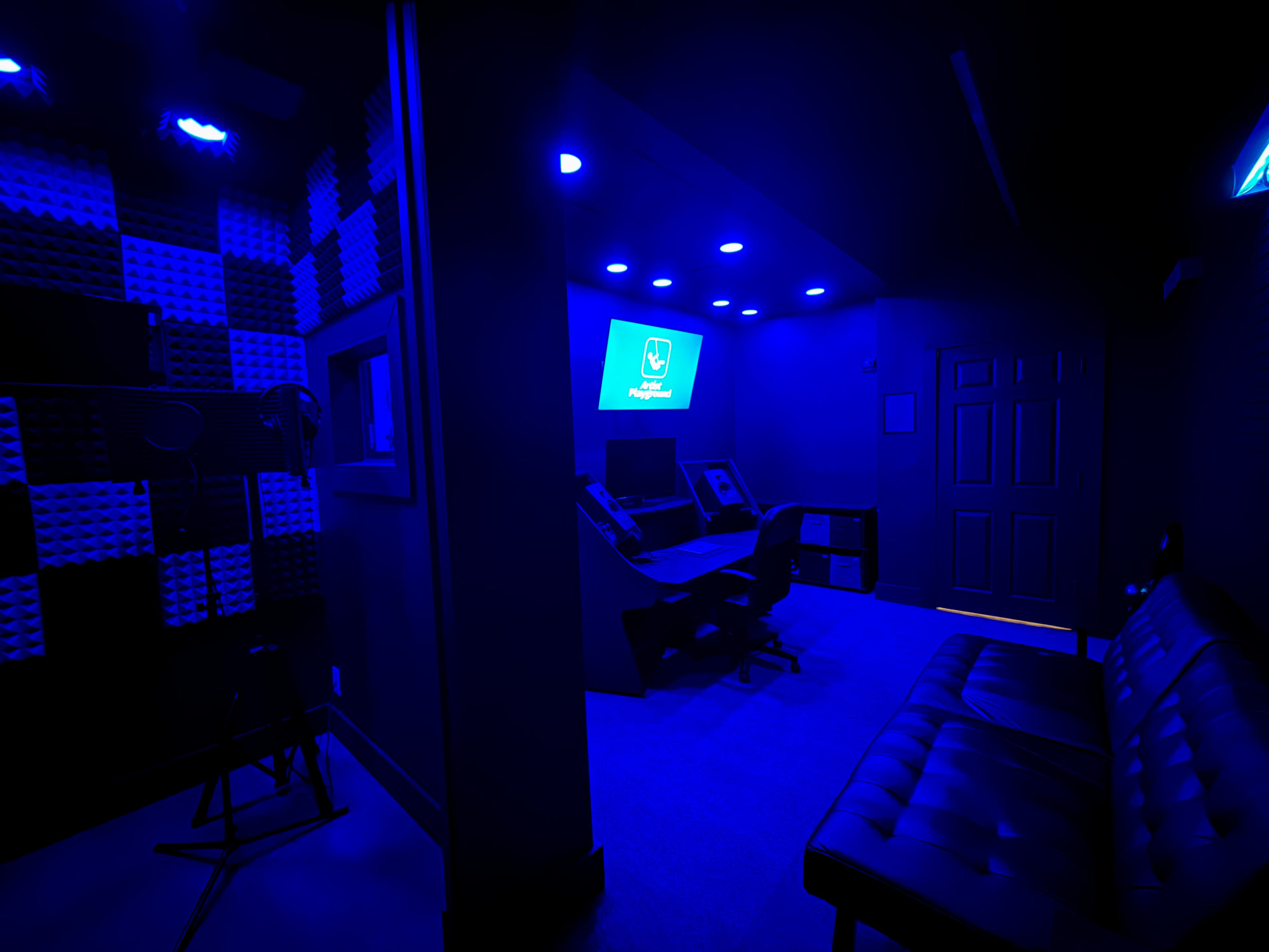 The image shows a dimly lit audio recording studio featuring a control desk, a seating area, and soundproof foam on the walls, all illuminated by blue lighting.