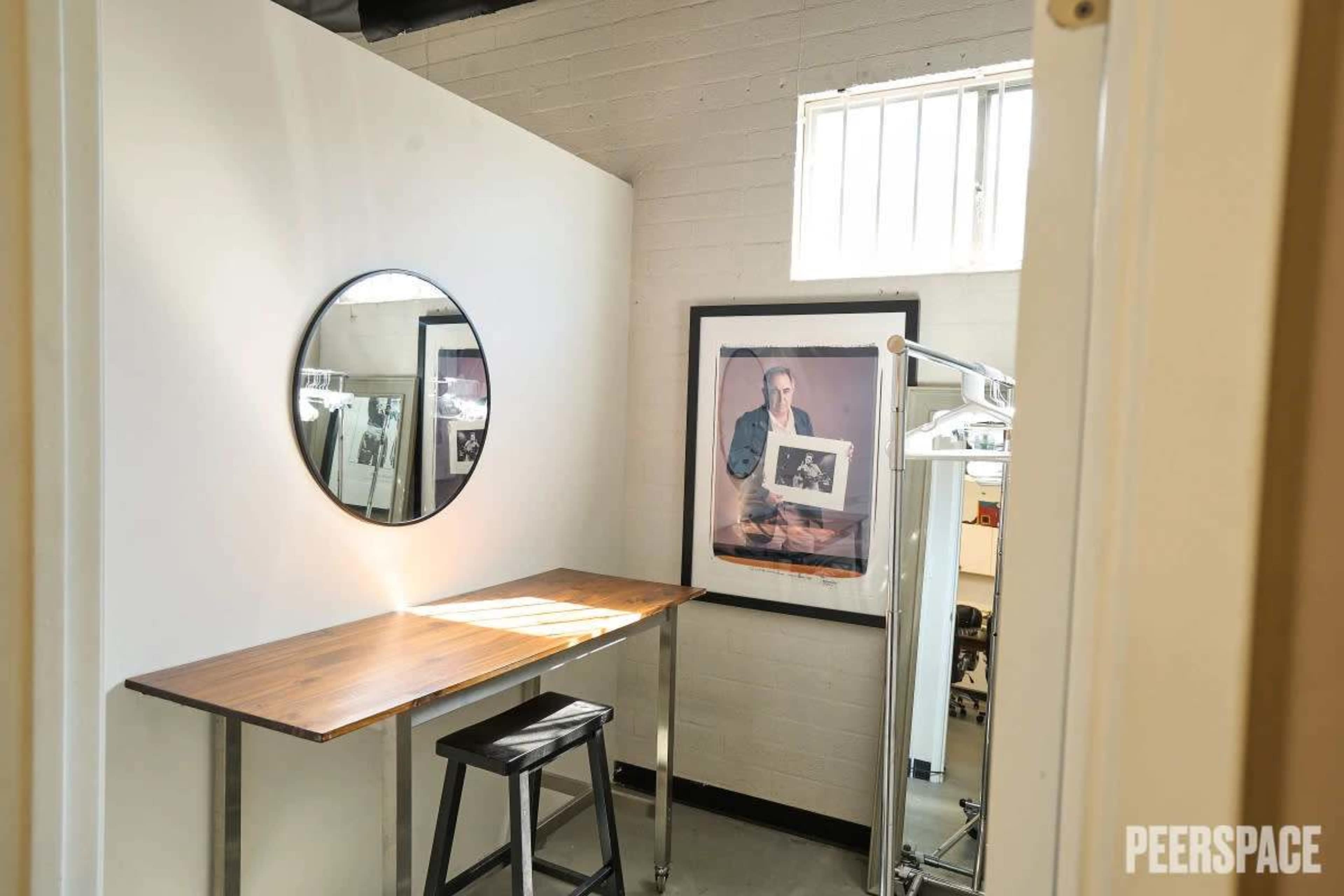 The image shows a small room featuring a wooden table, a round mirror on the wall, a black stool, and a framed picture of a man hanging nearby.