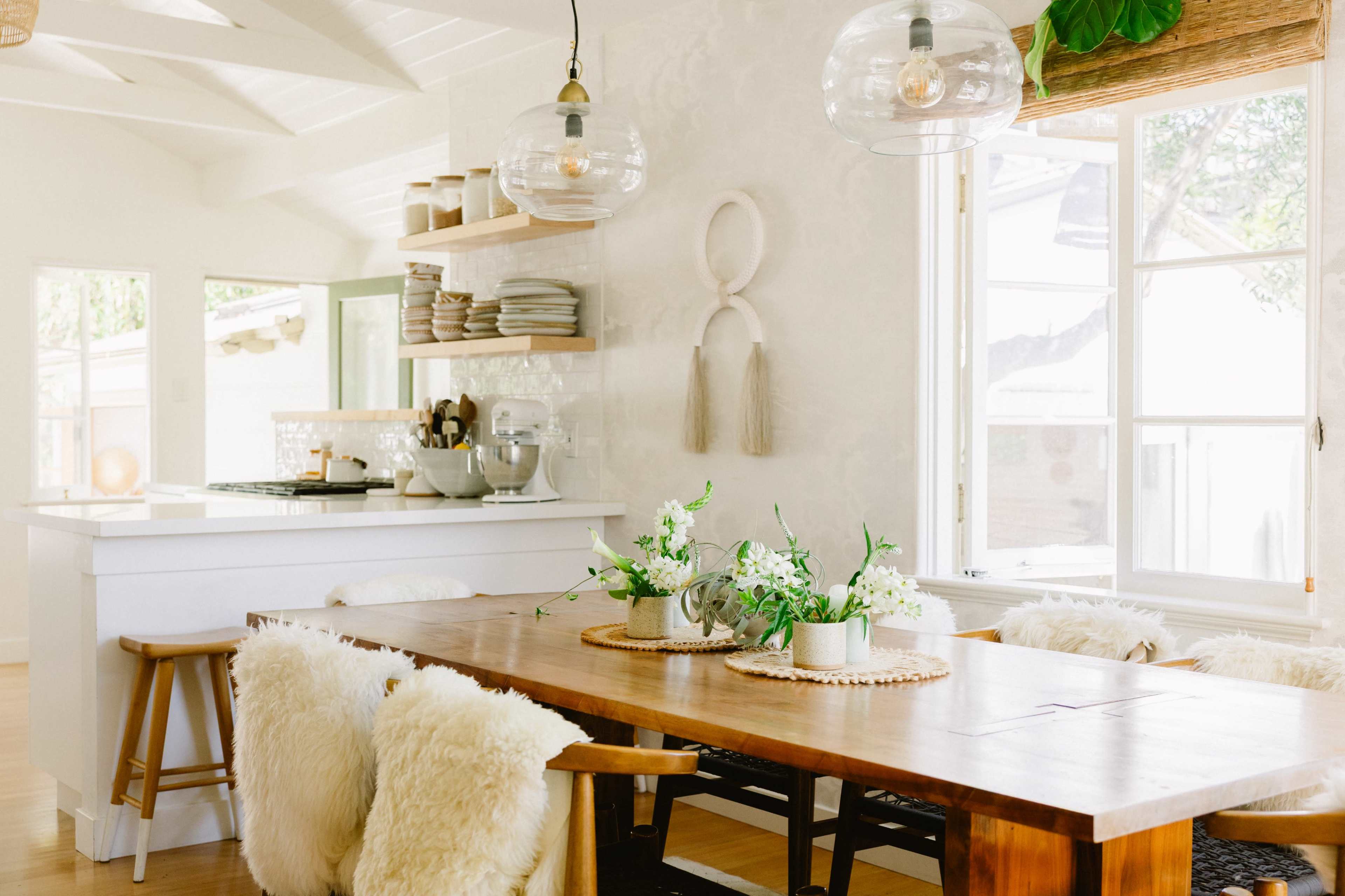 A dining area features a wooden table surrounded by cushioned chairs, with hanging pendant lights and a backdrop of a modern kitchen.