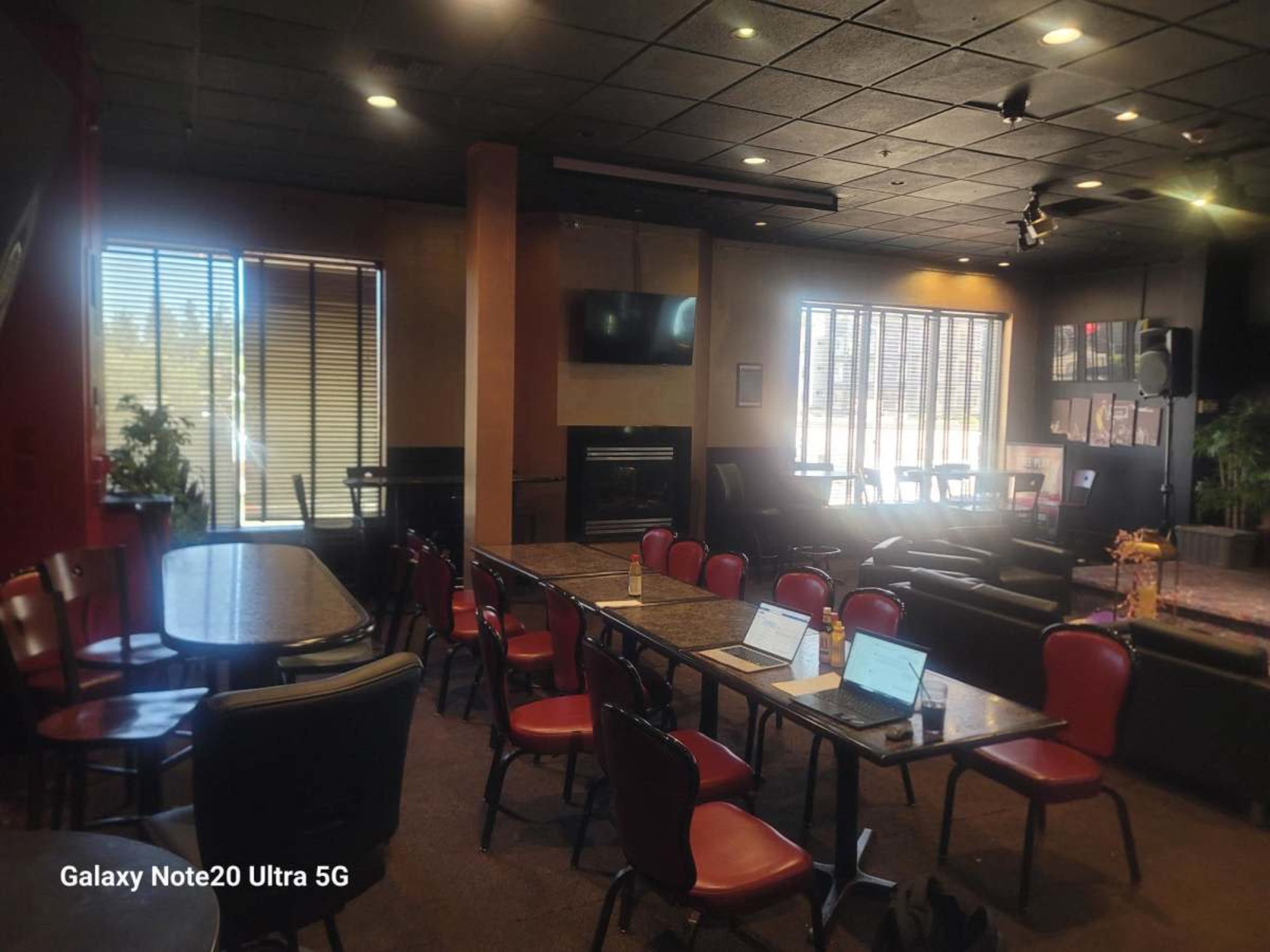The image shows a spacious interior of a cafe or meeting room with tables and chairs arranged neatly, a large TV on the wall, and a fireplace.