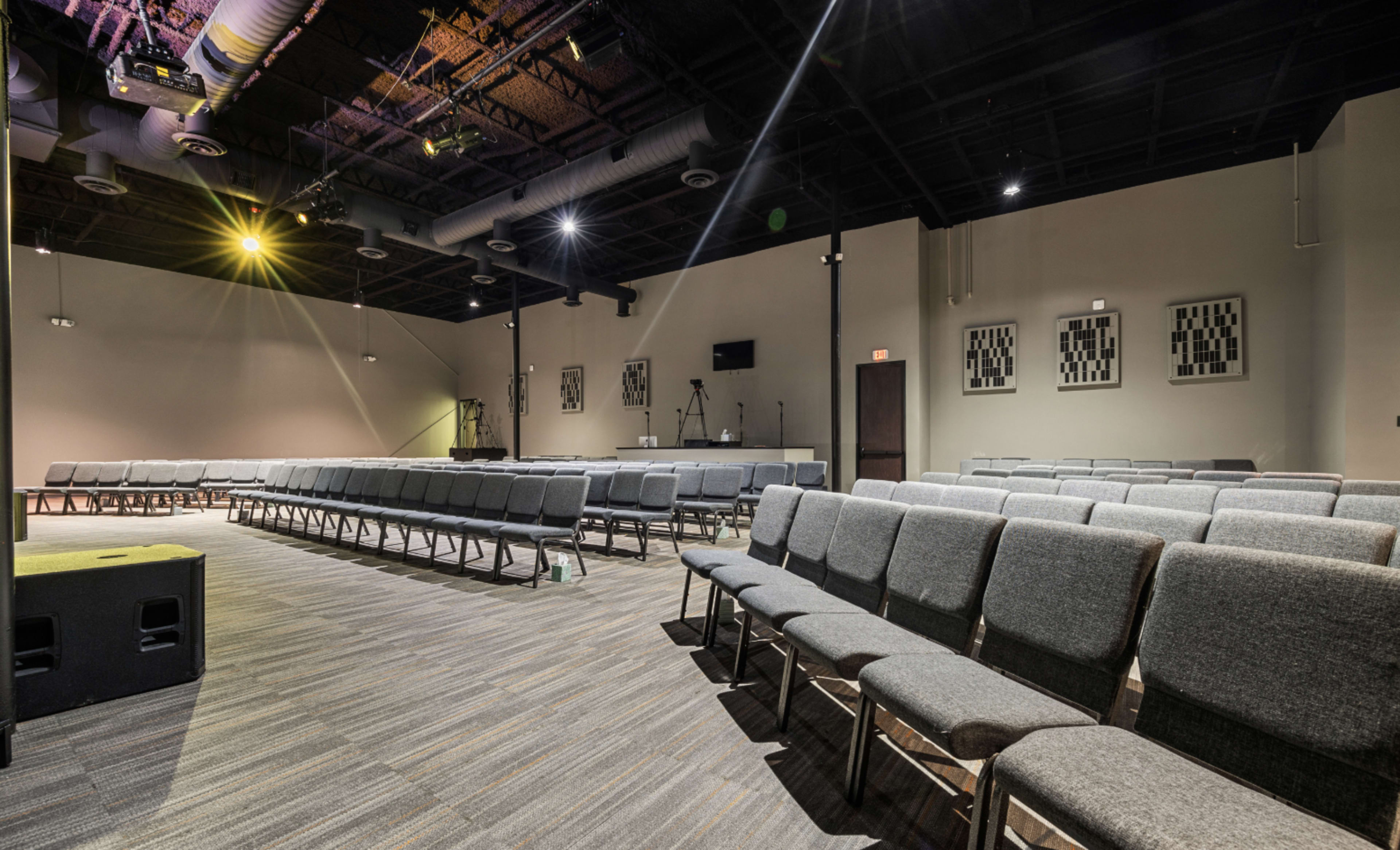 The image shows a modern auditorium with rows of gray chairs arranged facing a stage area, illuminated by overhead lights.