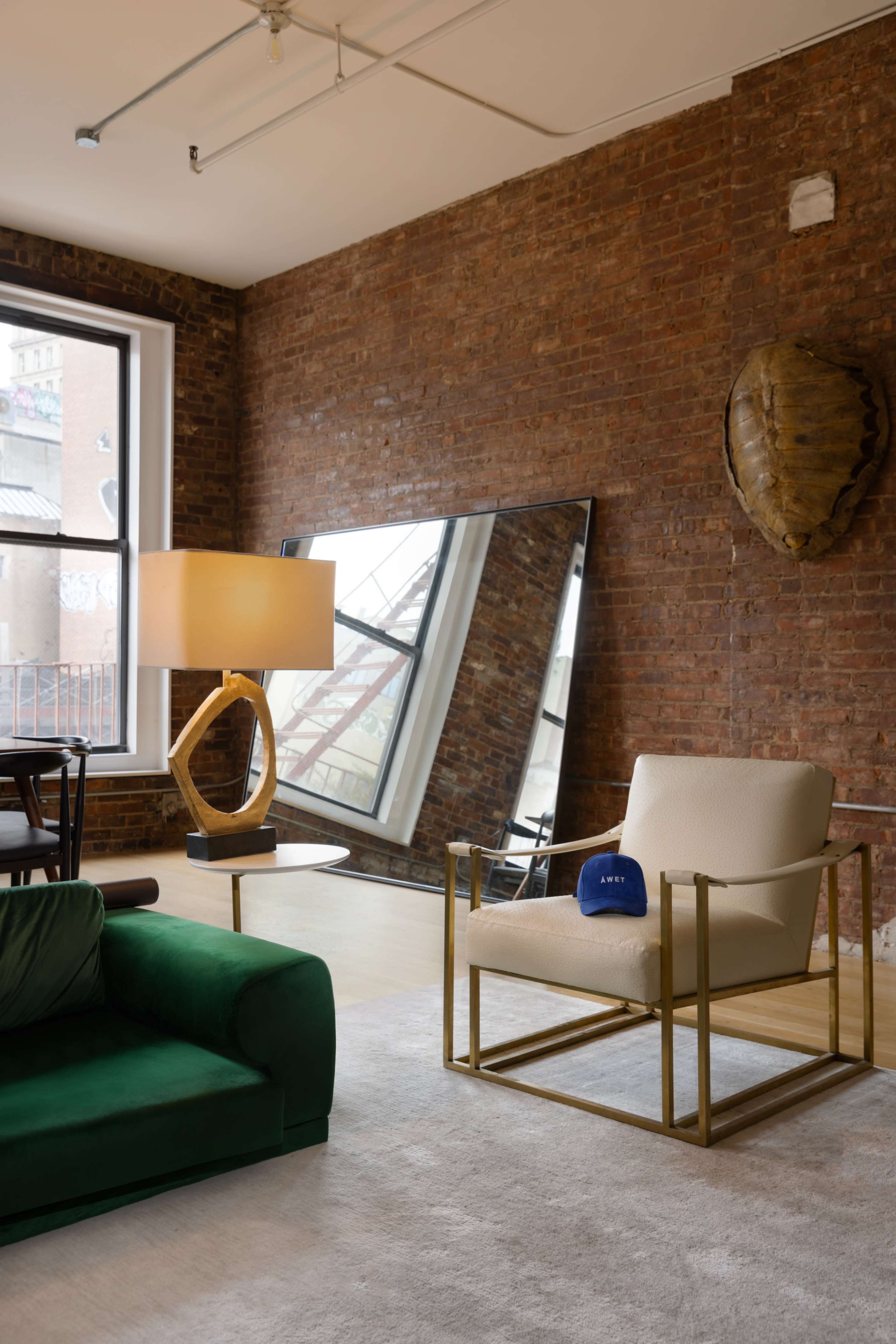 A modern living room features a green sofa, a beige armchair with gold metal framing, a large mirror reflecting a brick wall, and a floor lamp beside a circular side table.