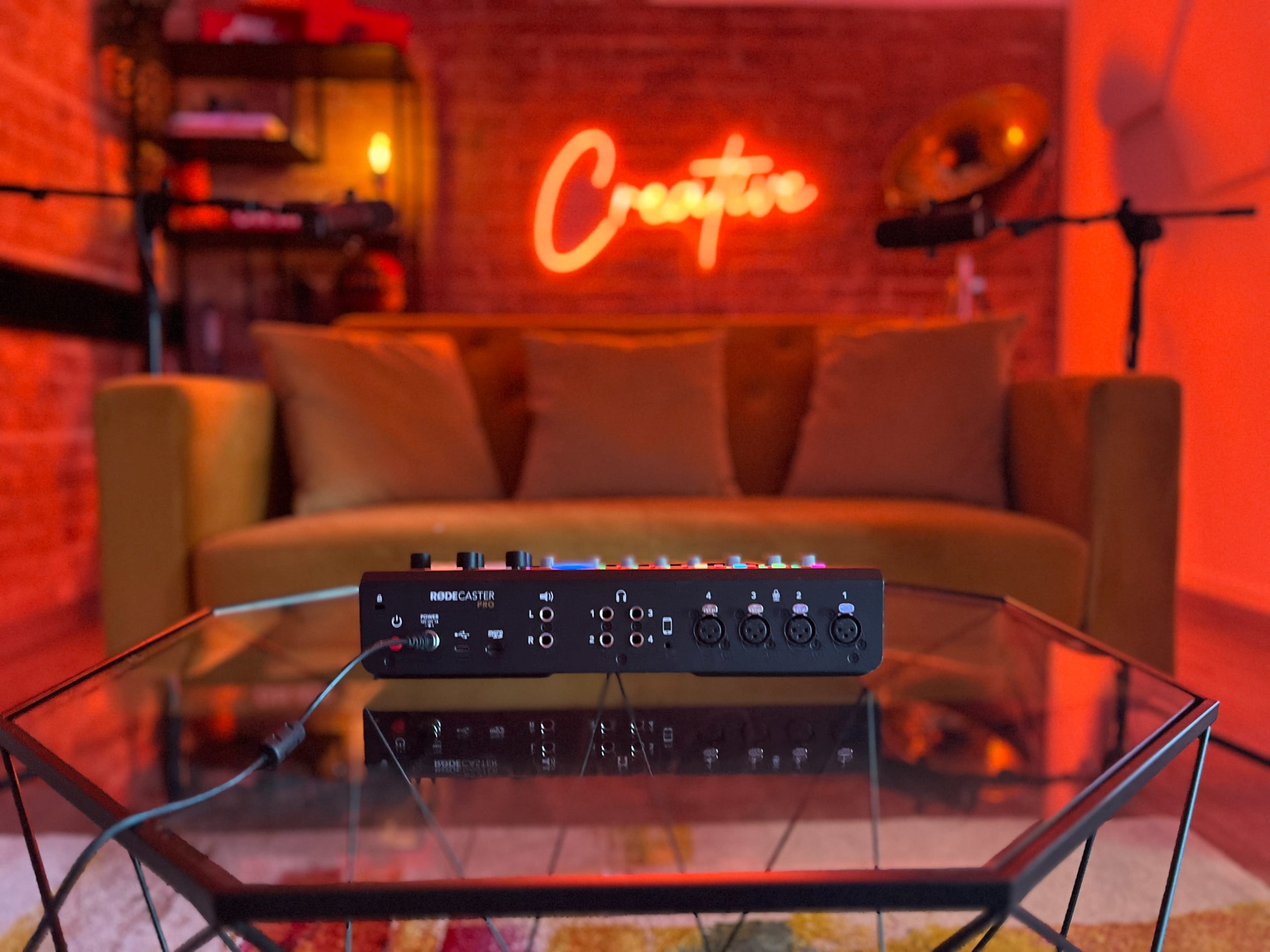 A music mixer placed on a glass table in a cozy room with a neon sign that reads "Creator" and a brown couch in the background.