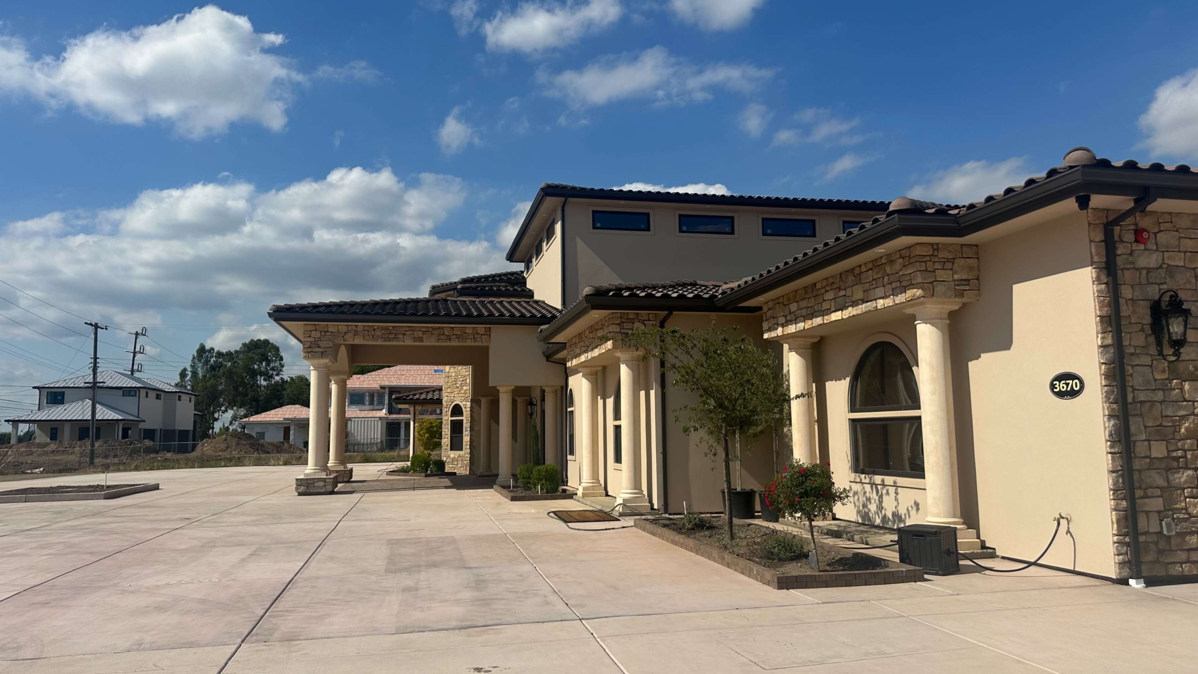 The image shows a modern, multi-story building with a combination of stone and stucco exterior, featuring columns and a landscaped entrance.