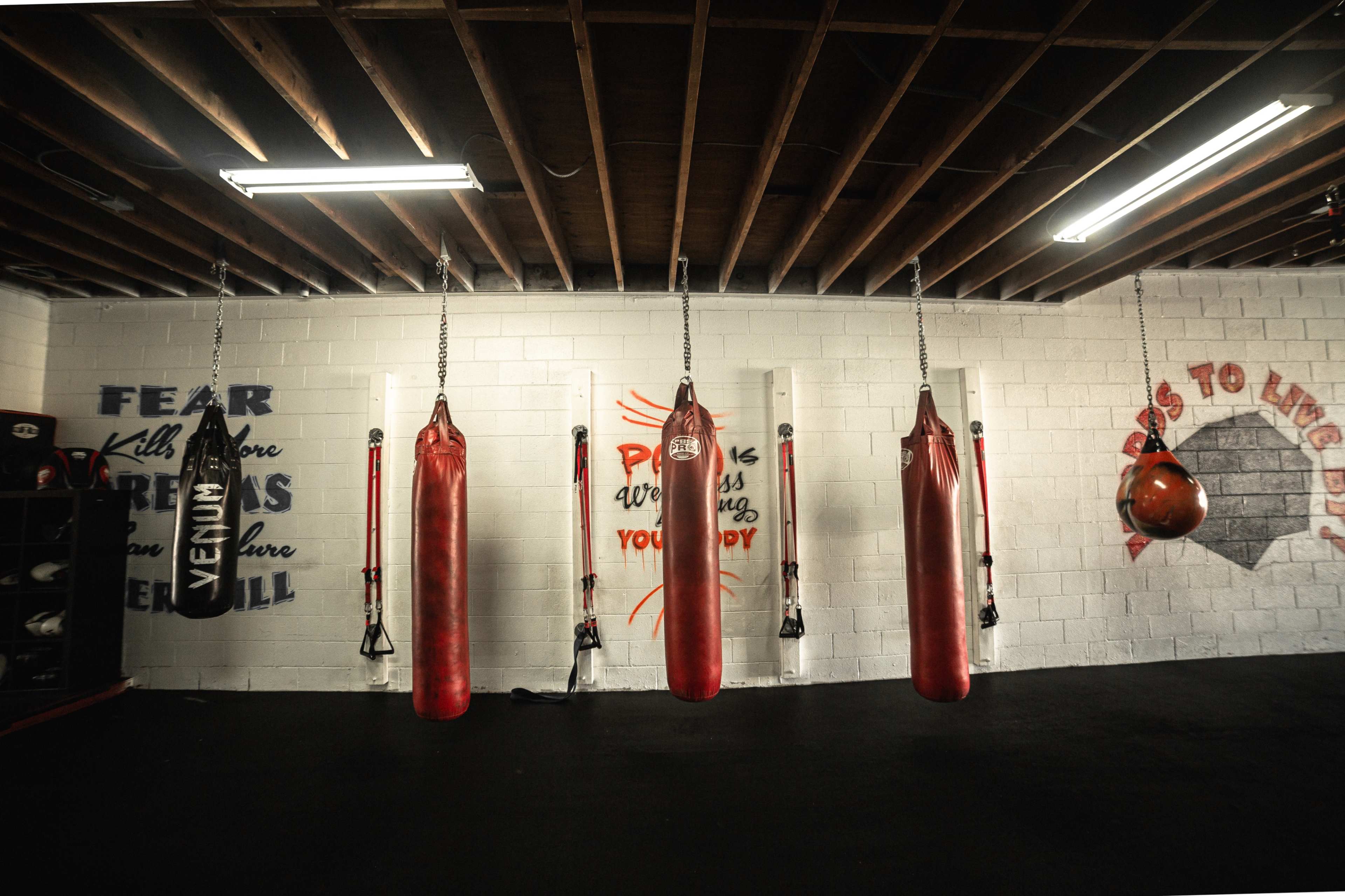 A boxing gym with several red heavy bags hanging from the ceiling, mounted on wall brackets, and motivational slogans painted on the wall.