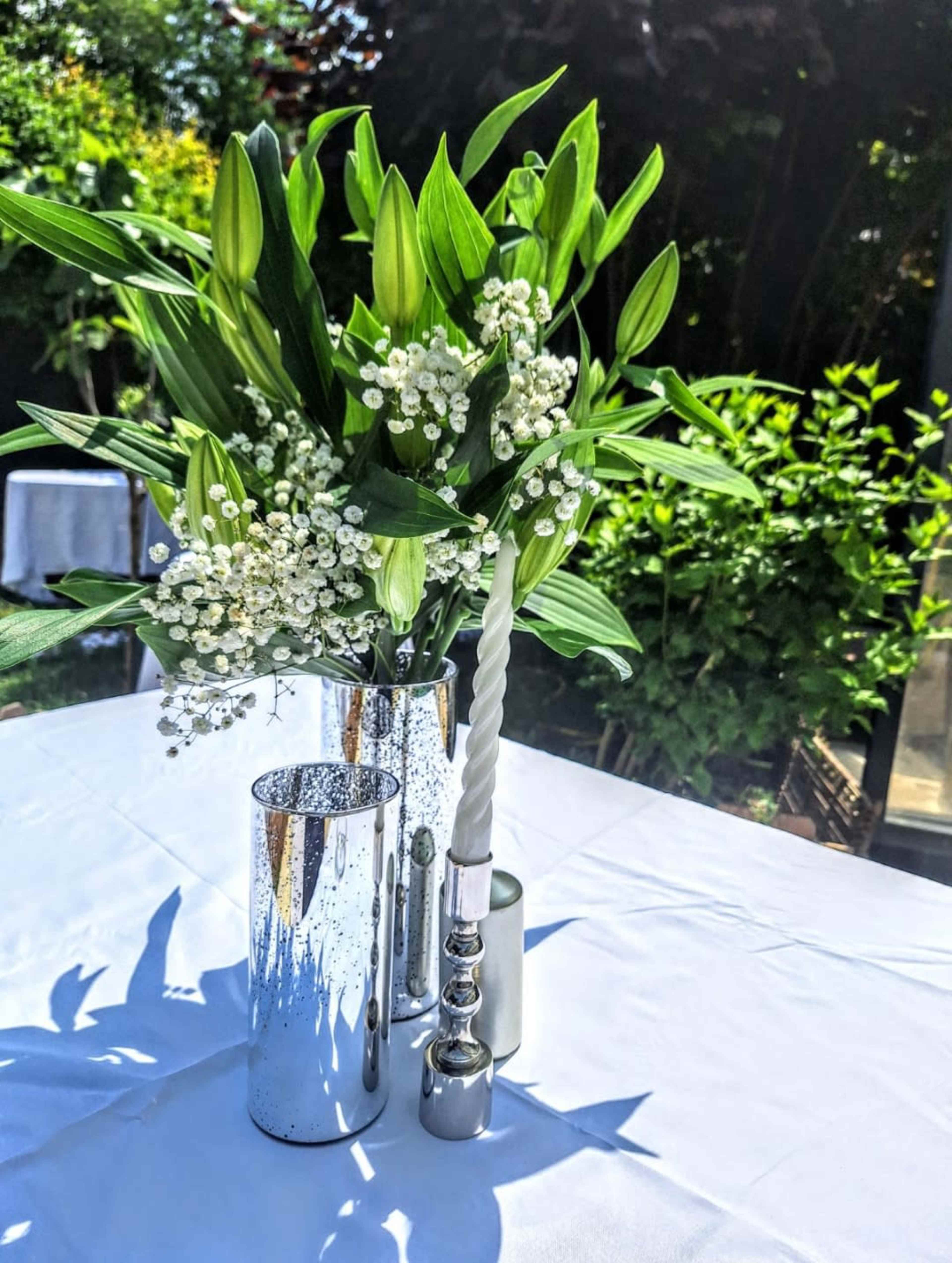 The image shows a table set with a twisted white candle and a silver vase containing white flowers, all surrounded by lush green foliage.