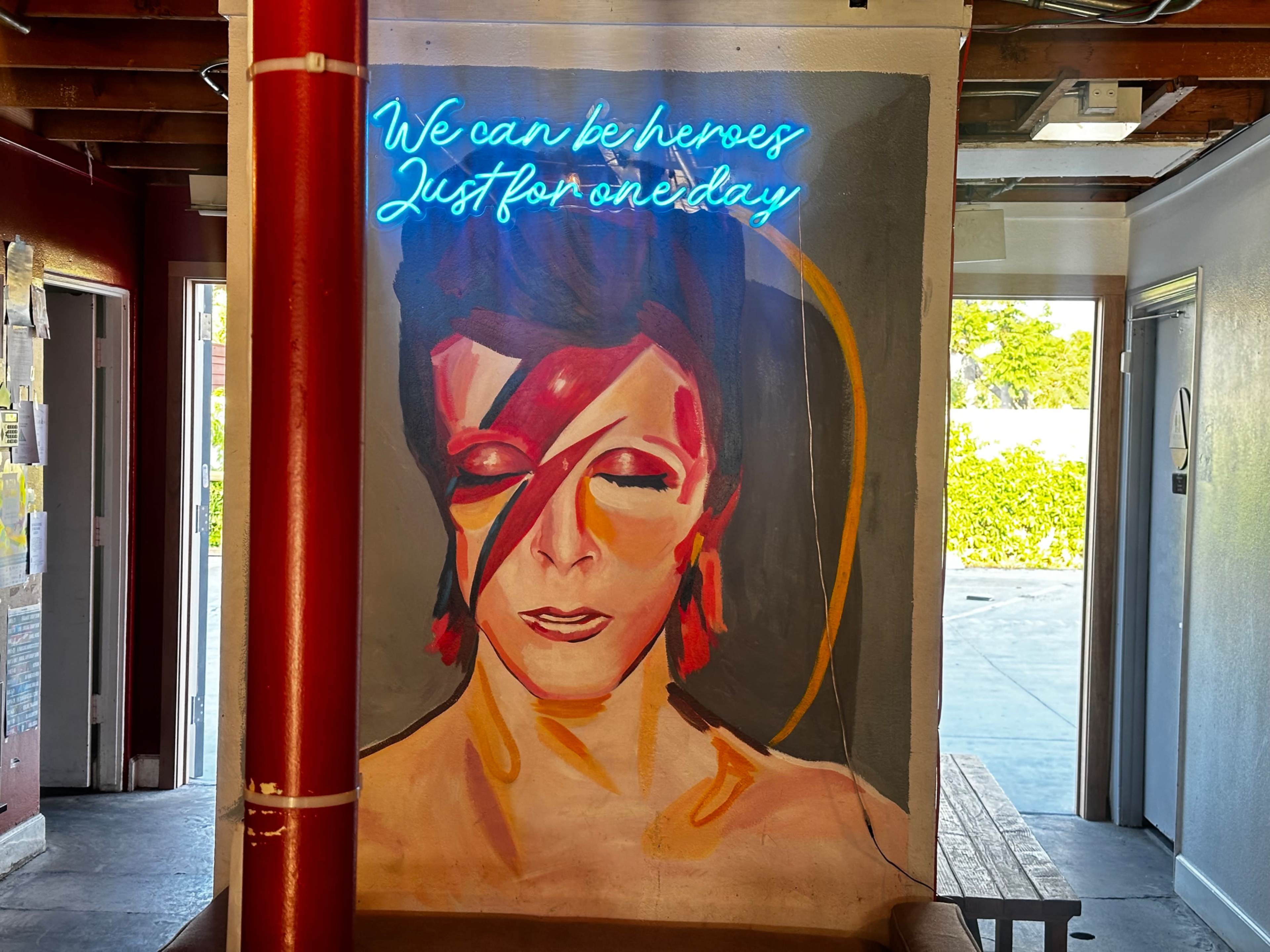 A colorful mural of a person with orange lightning bolt face paint is illuminated by a neon sign that reads, "We can be heroes, Just for one day."