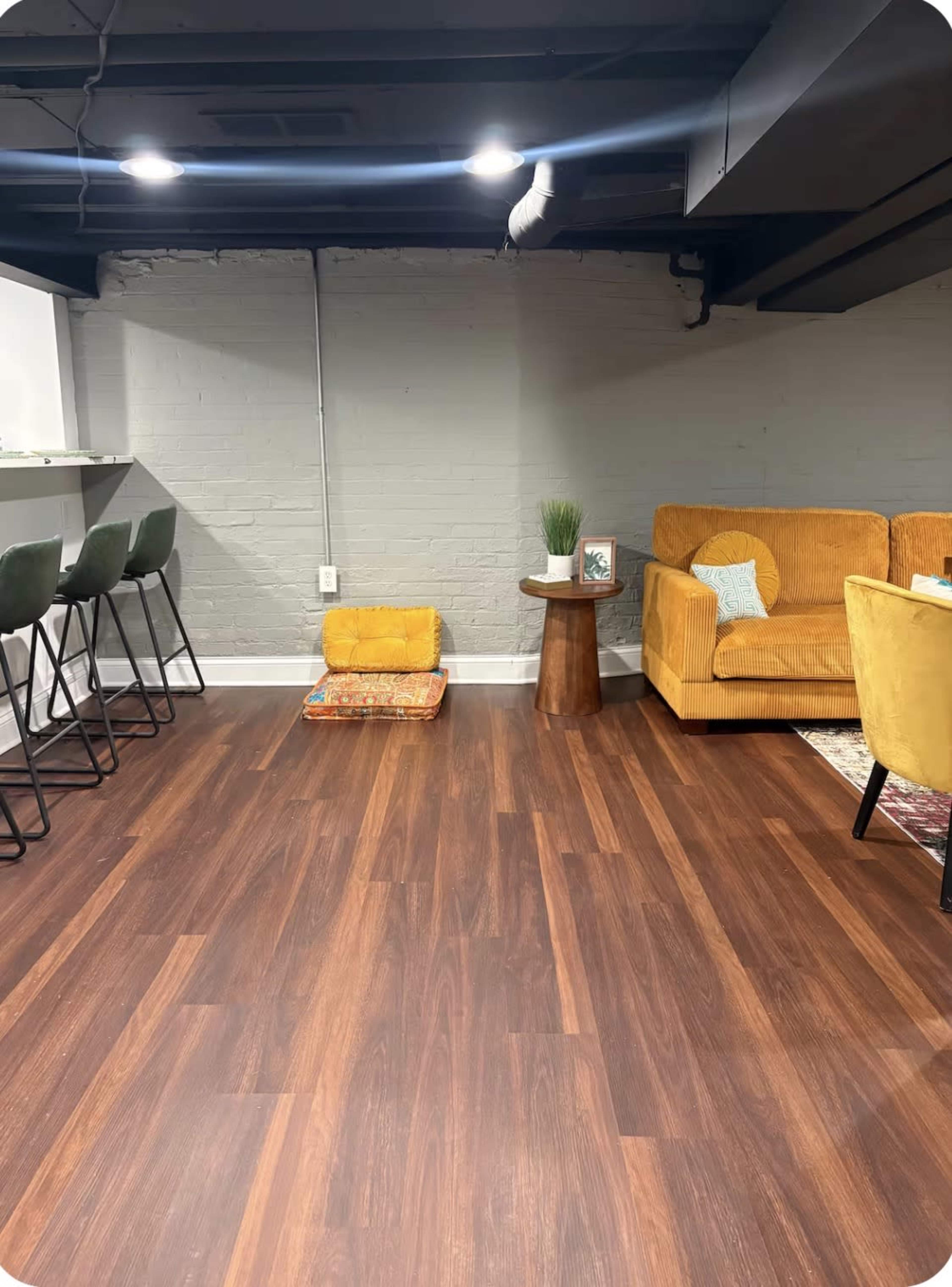 The image shows a modern basement living space featuring a yellow sofa, a small wooden table, bar stools, and a cozy area rug on the brown wooden floor.