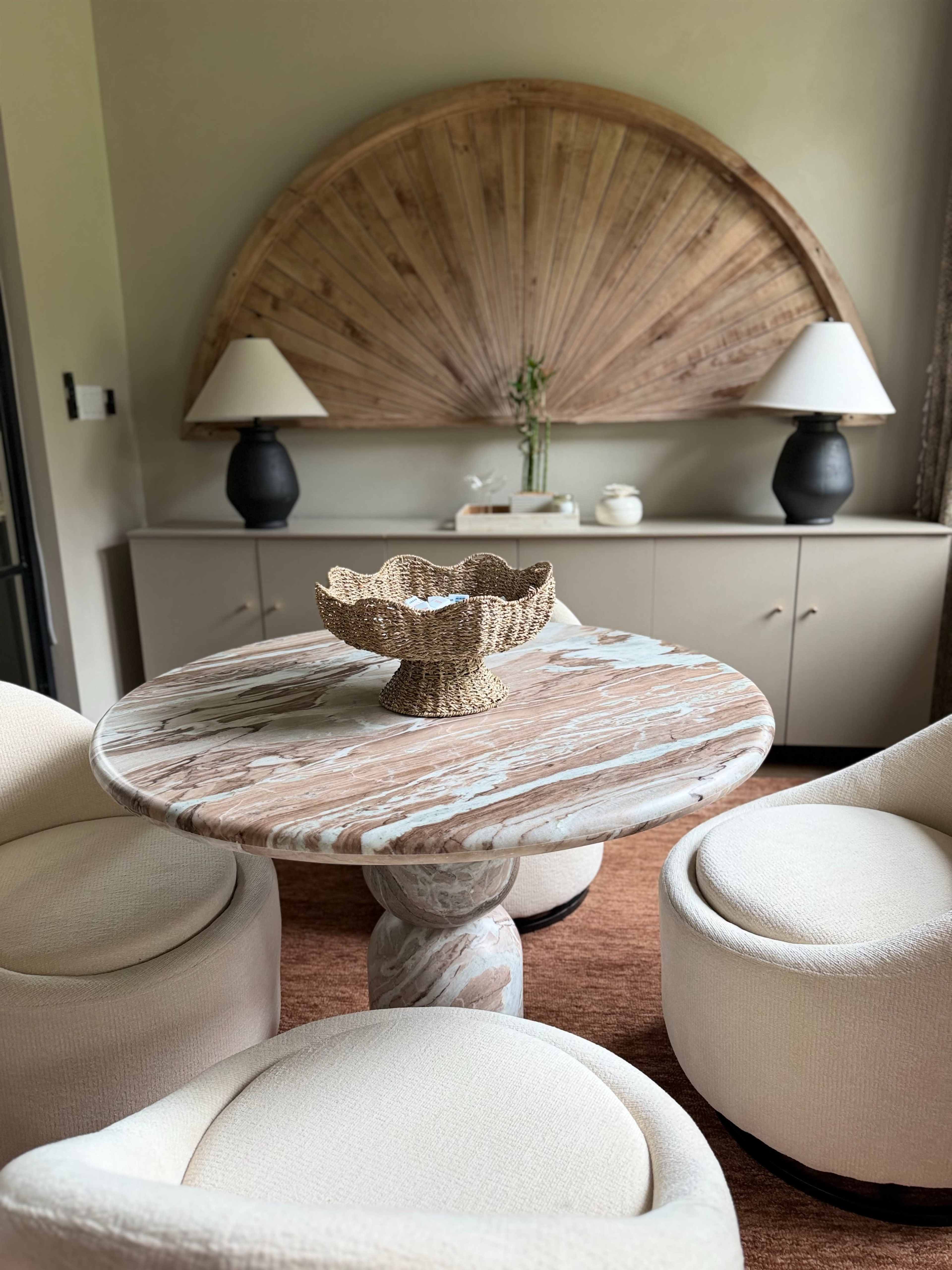 The image shows a circular marble table with a decorative bowl, flanked by two upholstered chairs, in a room adorned with a large wooden wall feature and two lamps on a sideboard.