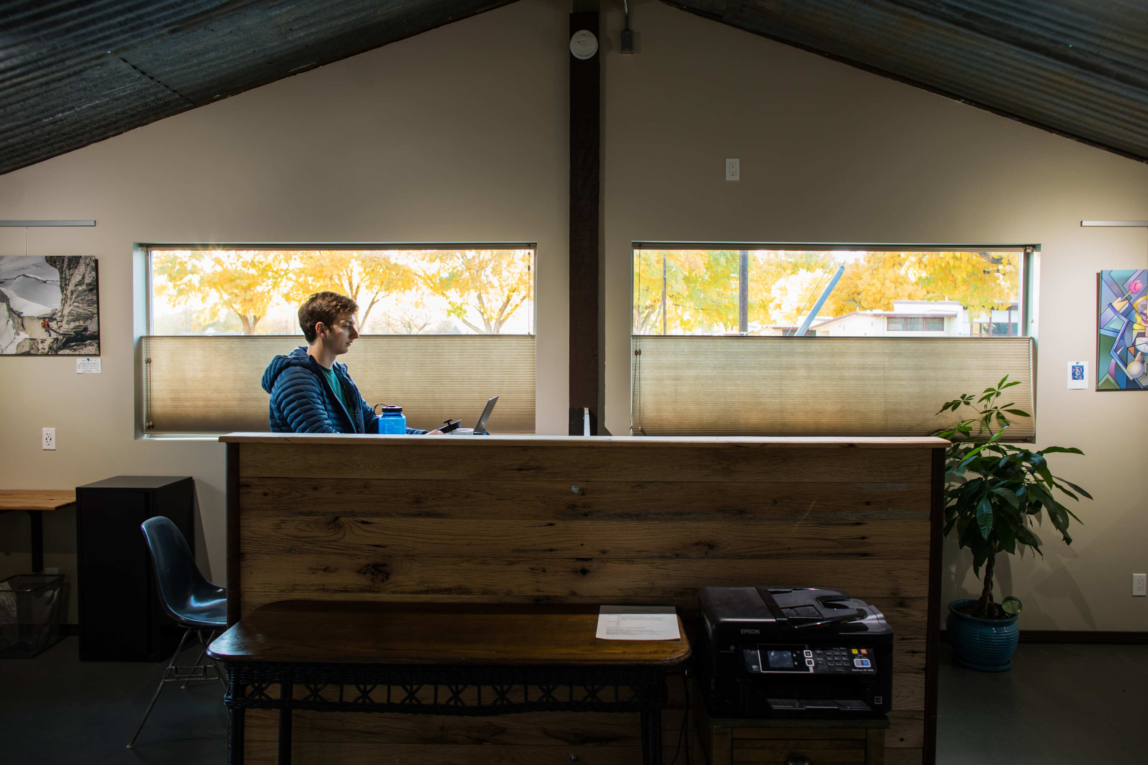 A person sits at a desk in an office space with large windows displaying autumn foliage outside.