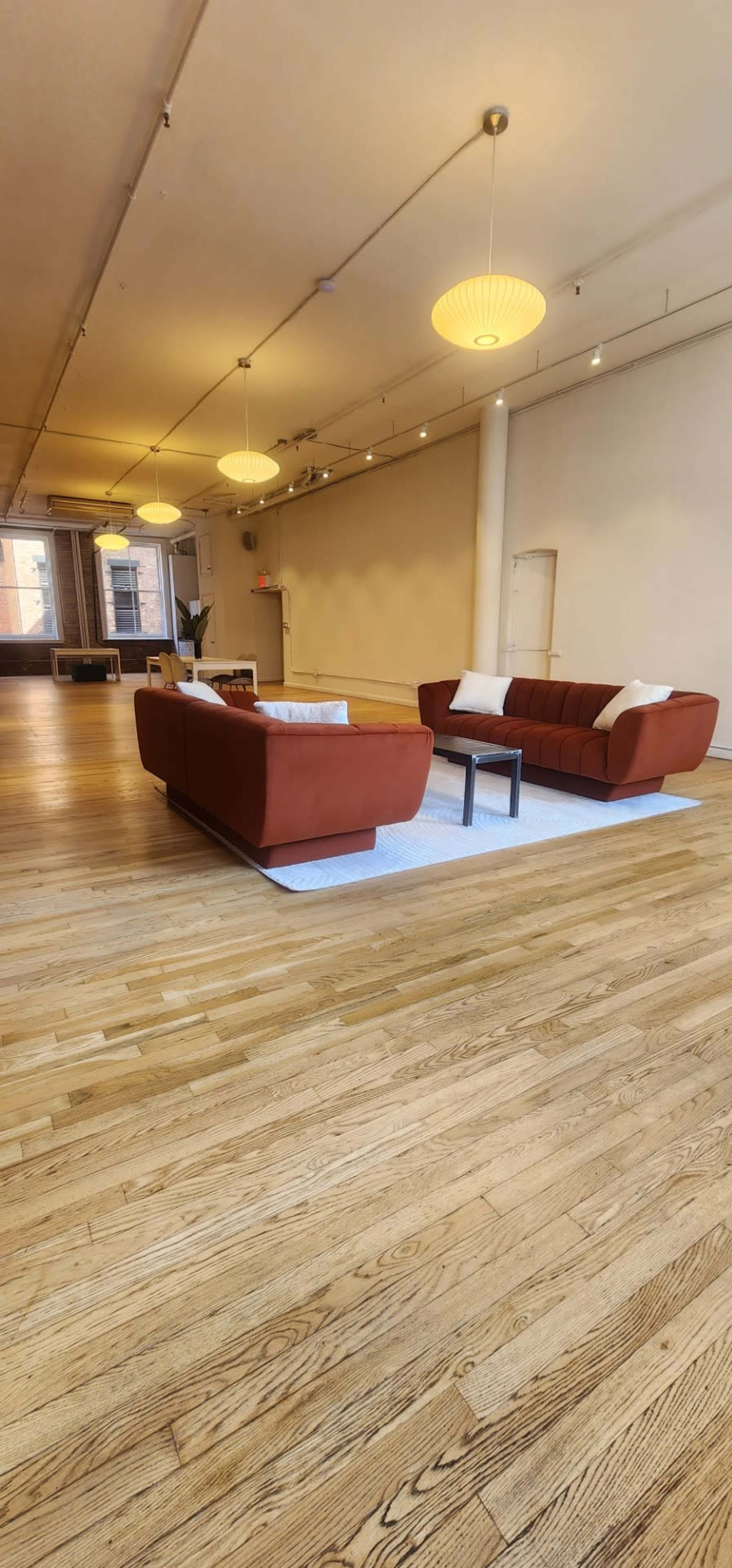 The image shows a spacious, minimalist room featuring two modern red sofas, a small table between them, and a wooden floor illuminated by pendant lights.