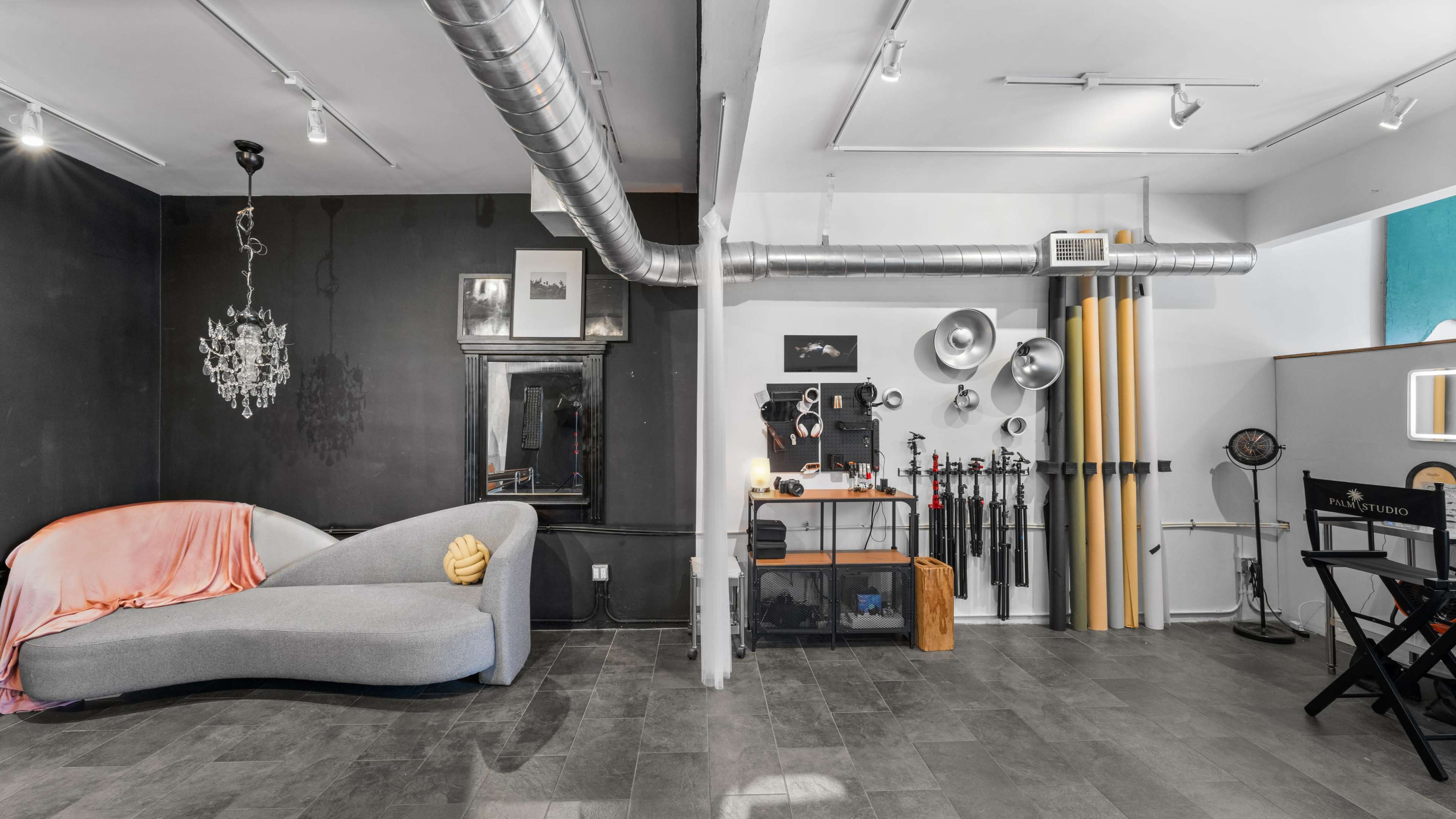 The image shows a stylish studio space with a gray sofa, black walls, and various photography equipment displayed along one side.