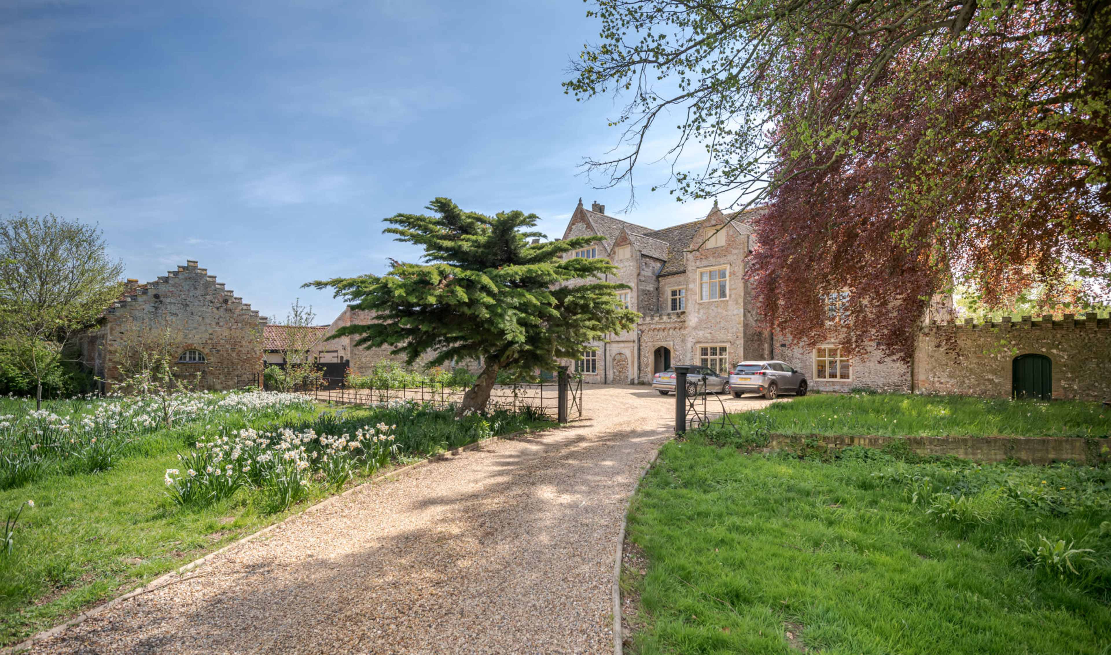 A gravel path leads to a large stone house surrounded by greenery and flowering plants.