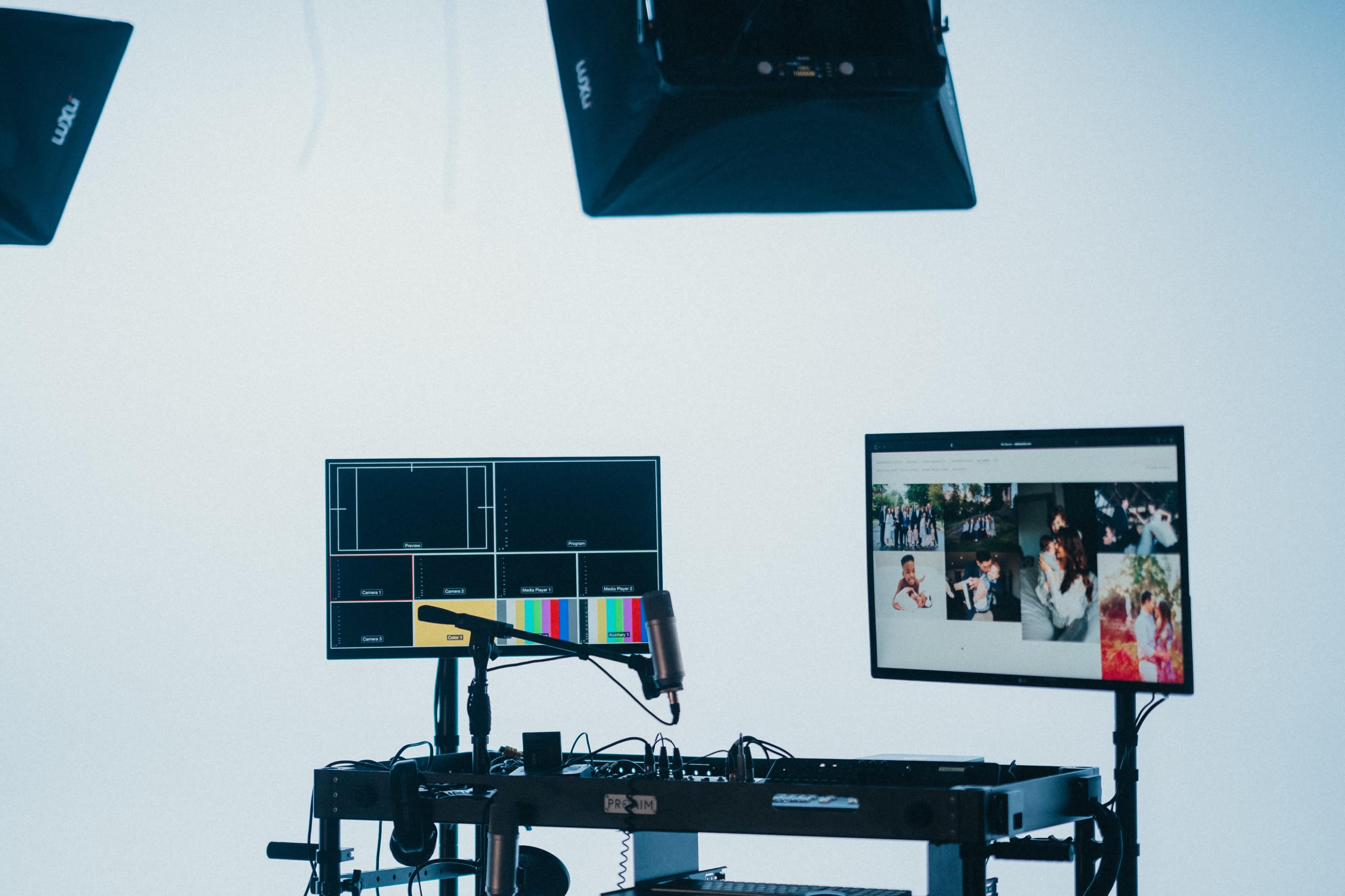 The image shows a studio setup with two computer monitors, a microphone, and studio lights against a plain background.