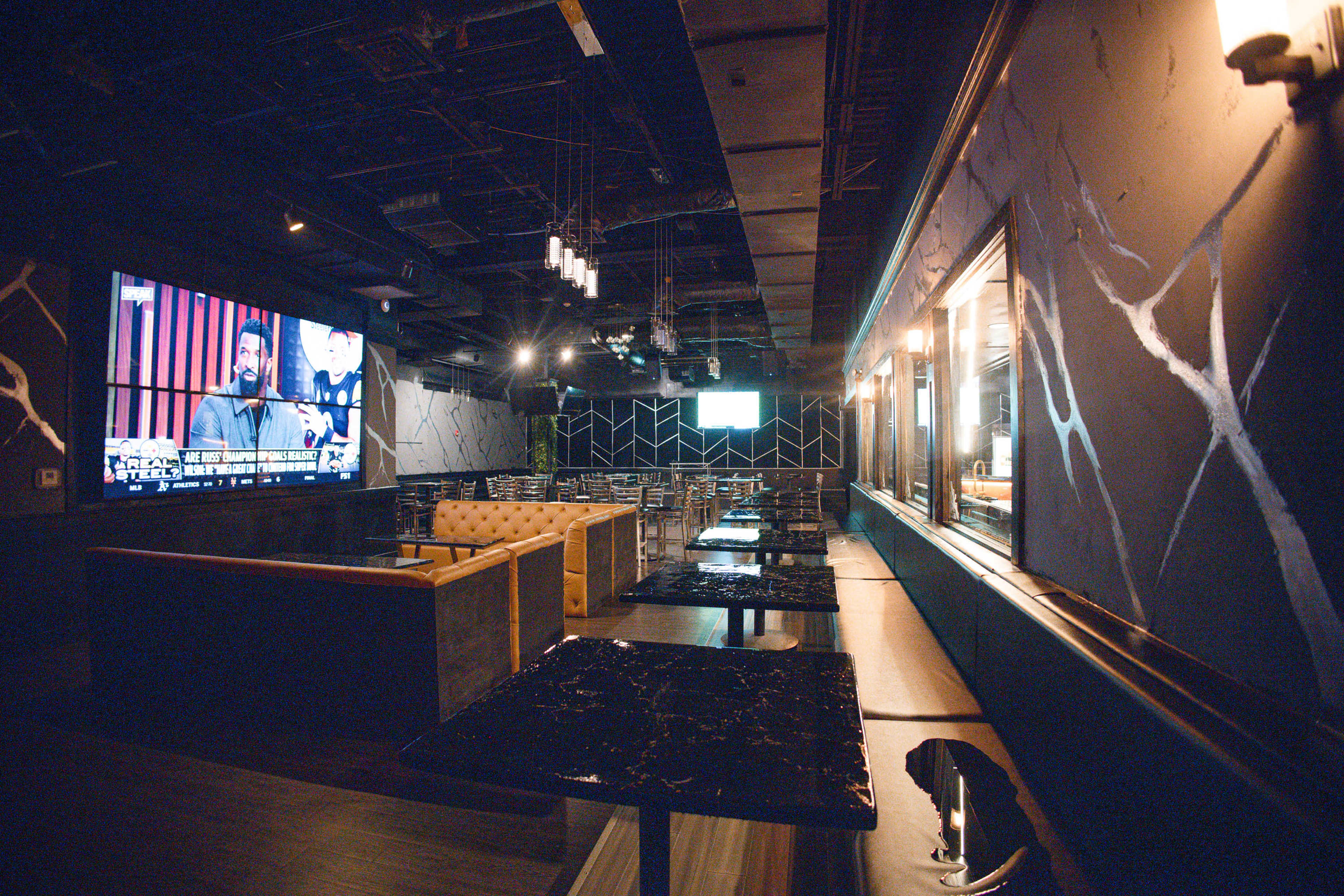 The image shows a modern lounge or bar interior with sleek black tables, plush seating, and large screens displaying content on the walls.