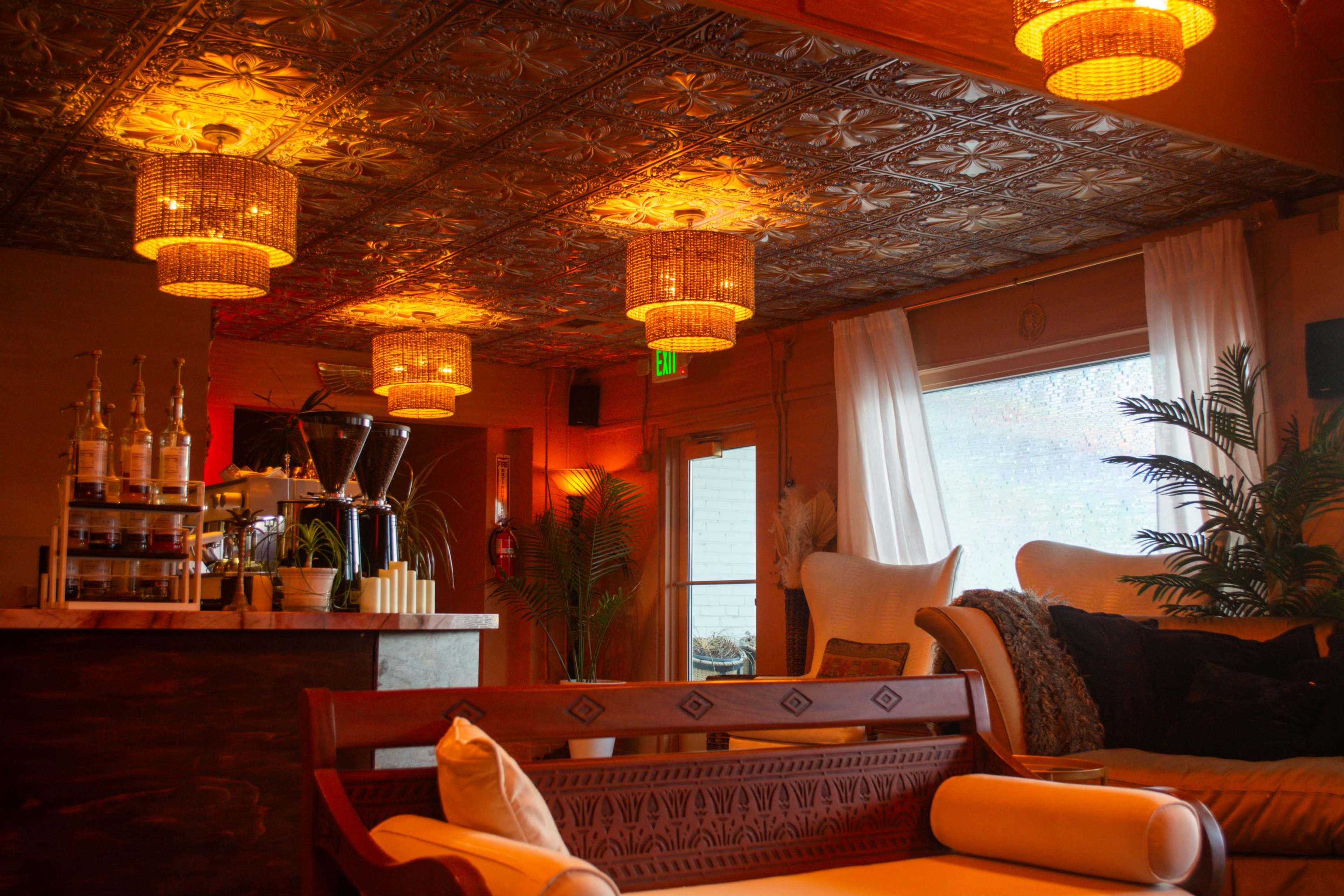 The image shows a cozy lounge area featuring a bar with bottles displayed, warm overhead lights, and comfortable seating next to large windows.