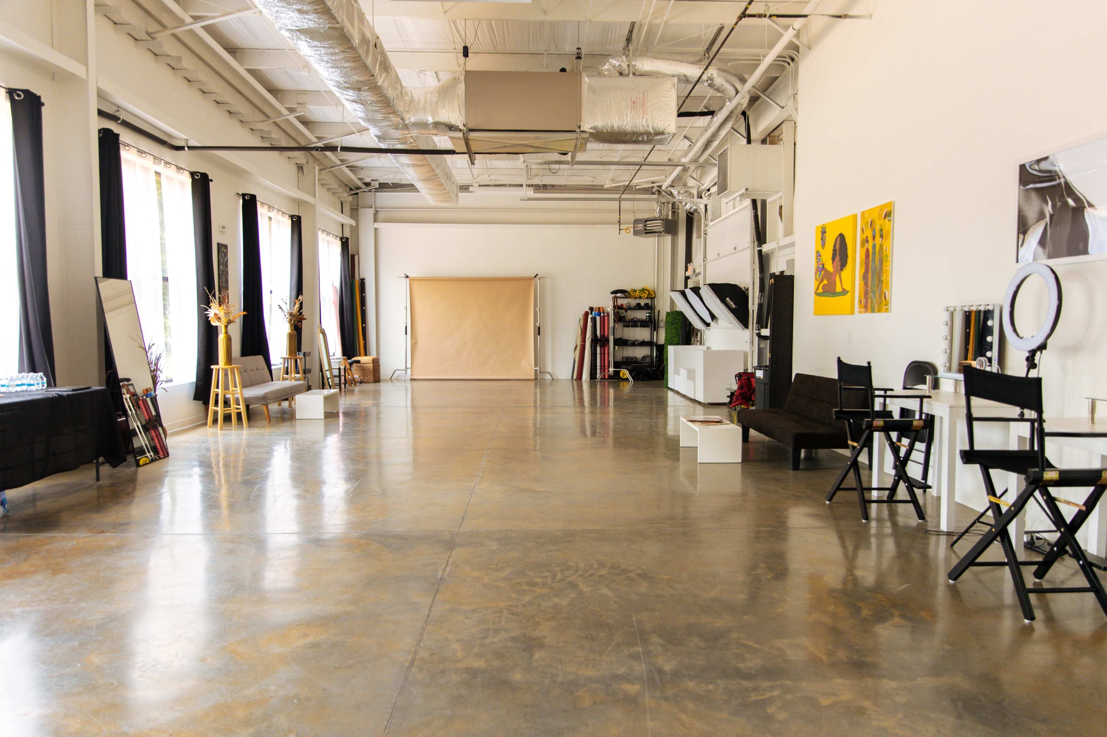 The interior of a spacious, well-lit studio features a polished concrete floor, large windows with sheer curtains, and various furniture pieces including a couch and director's chairs.