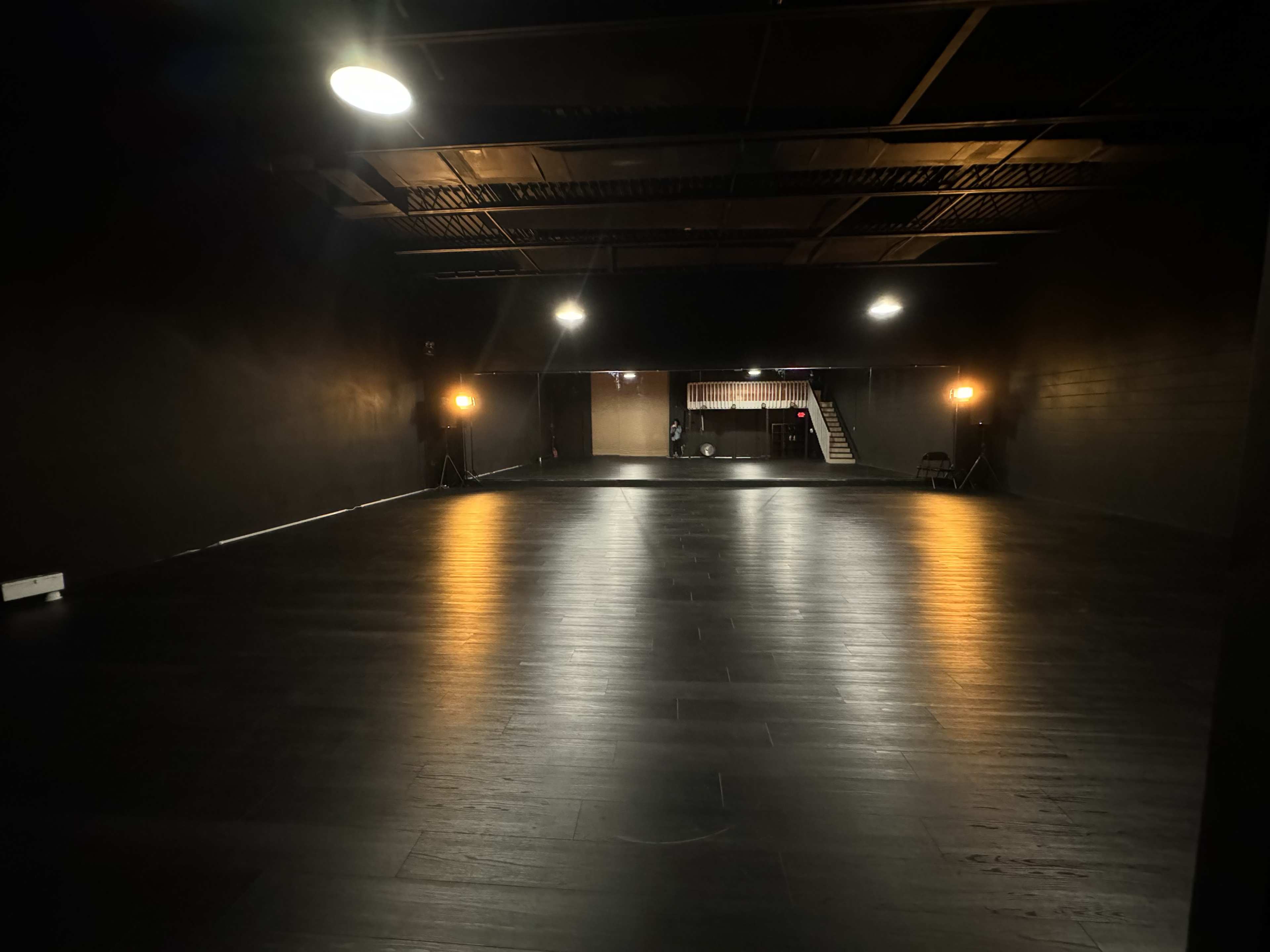 The image shows a spacious, dark room with a polished wooden floor, illuminated by lights positioned in two corners.