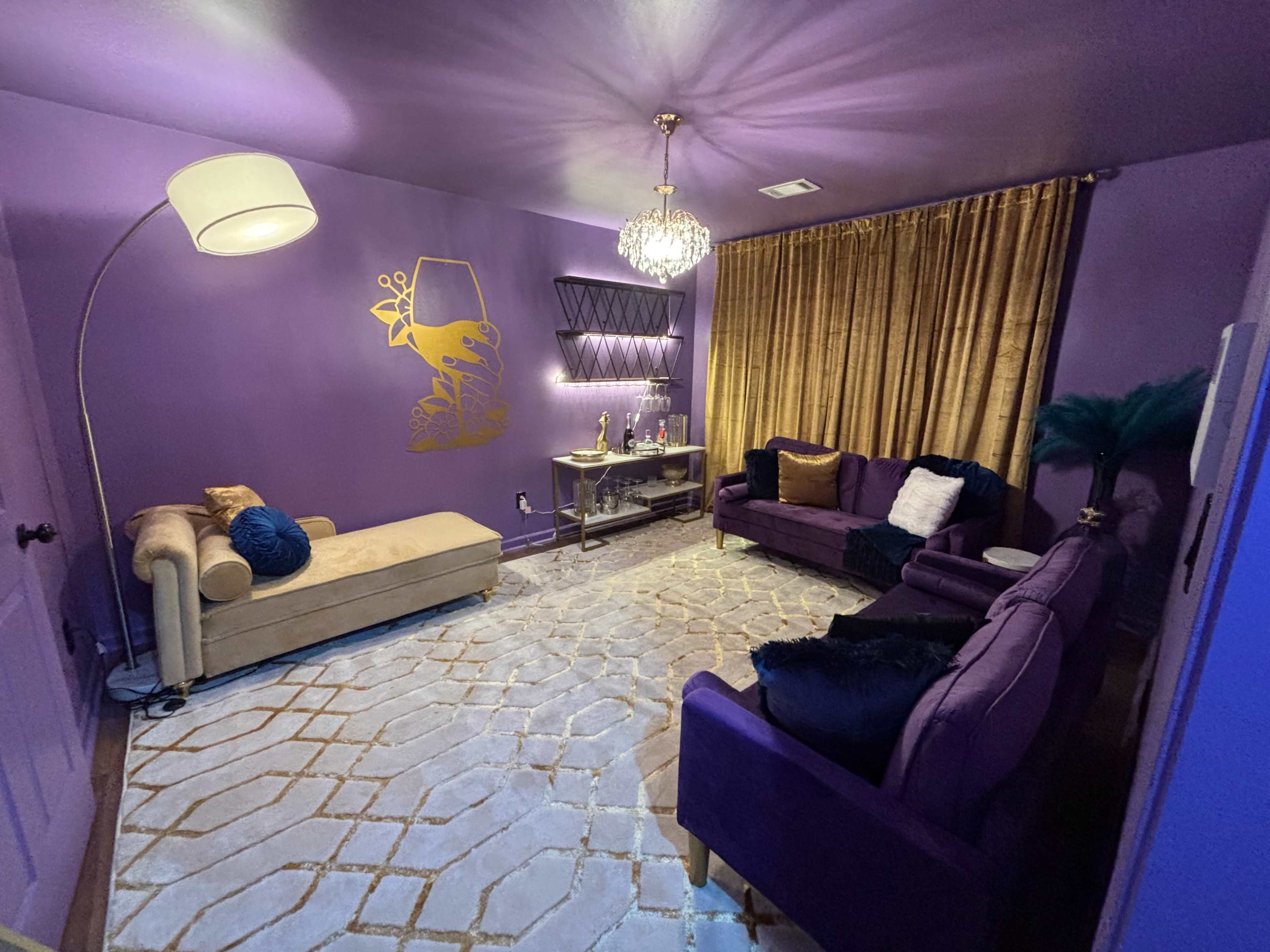 The image shows a purple-themed living room with a chaise lounge, a coffee table, two sofas, a large rug, and gold curtains.