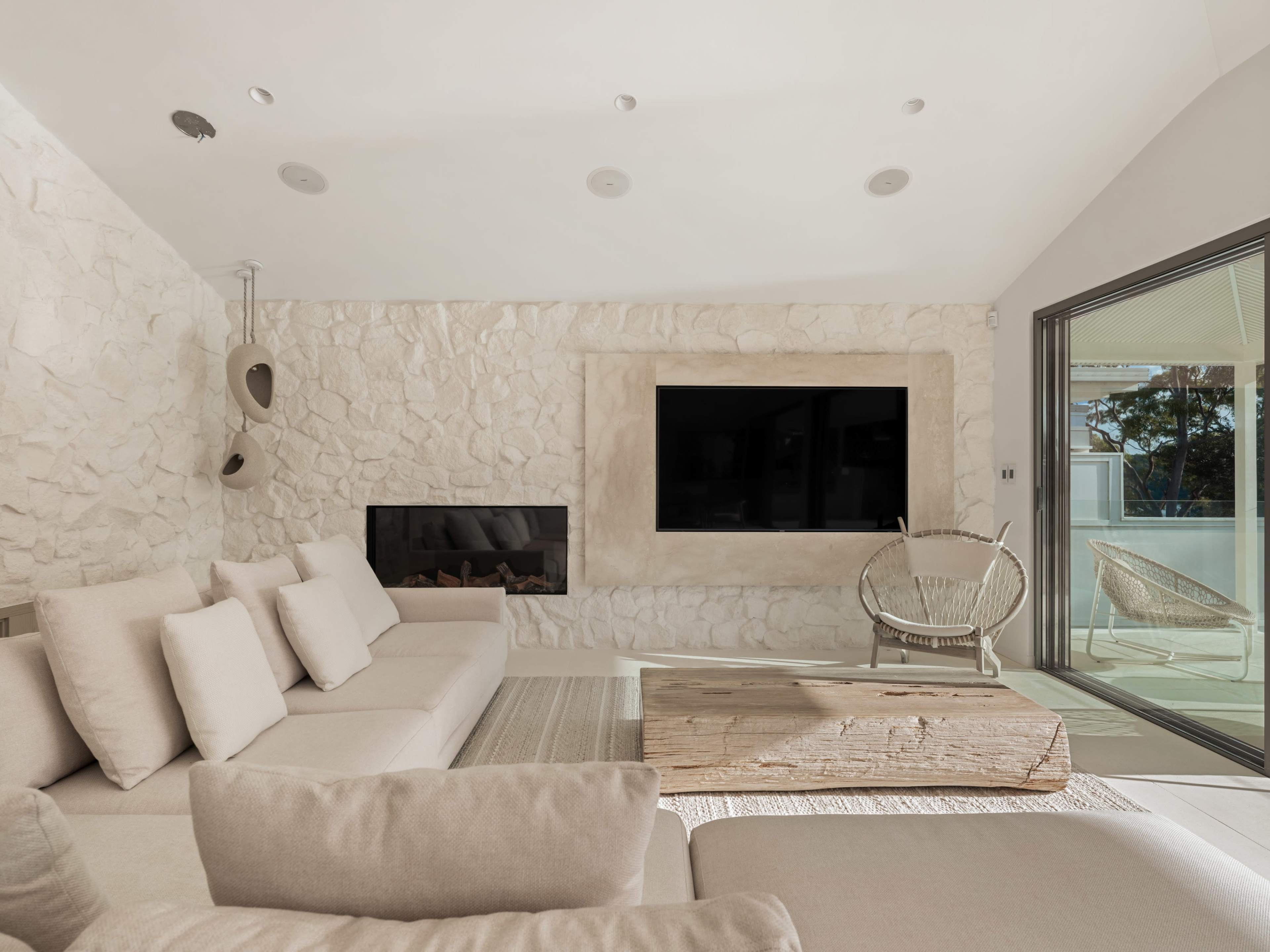 The living room features a light-colored stone wall, a large sectional sofa with multiple cushions, a wooden coffee table, and a flat-screen TV mounted above a fireplace.
