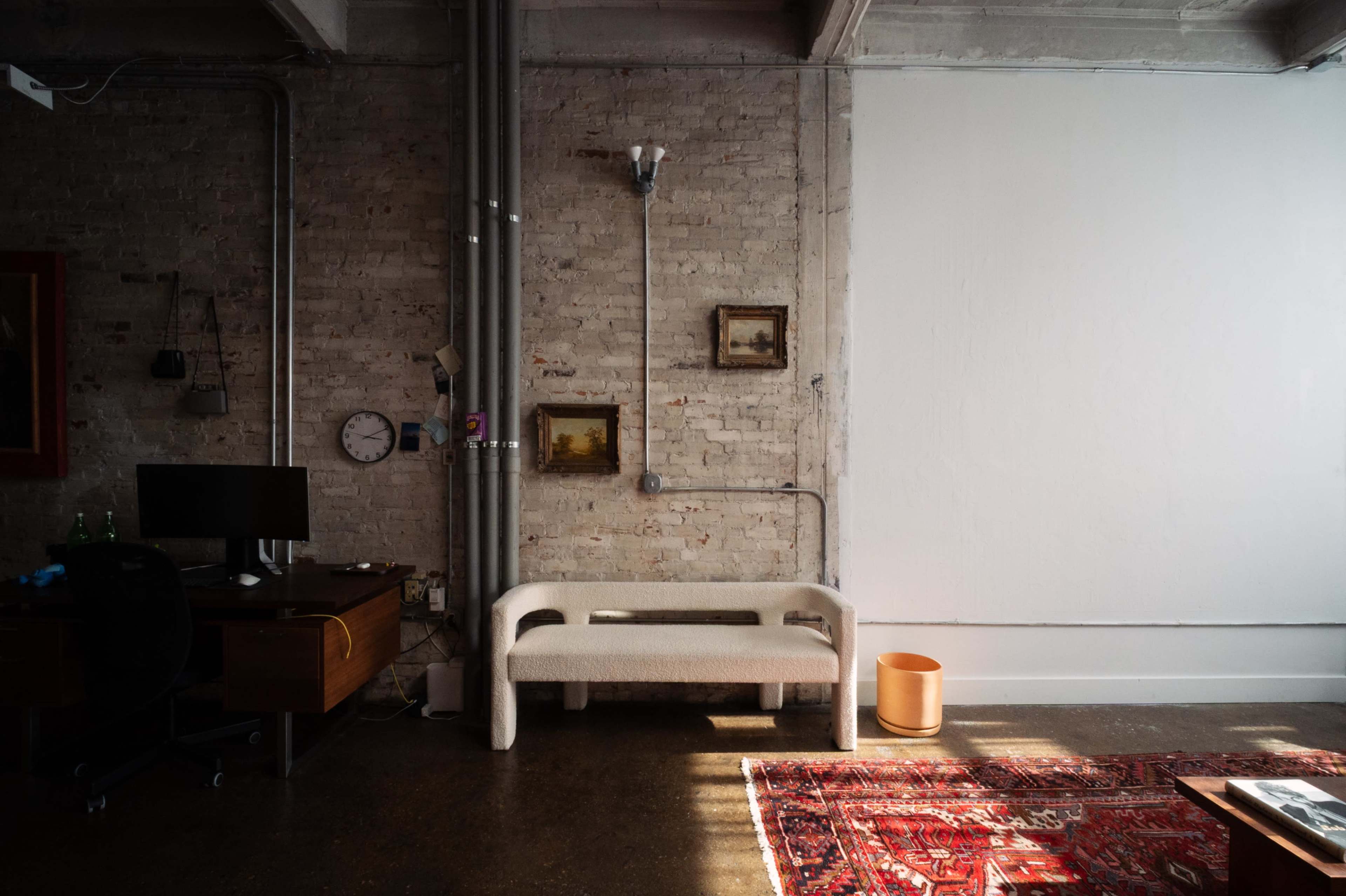 A modern interior features a minimalist couch, a desk with a computer, and a patterned rug on a concrete floor against a backdrop of exposed brick walls.