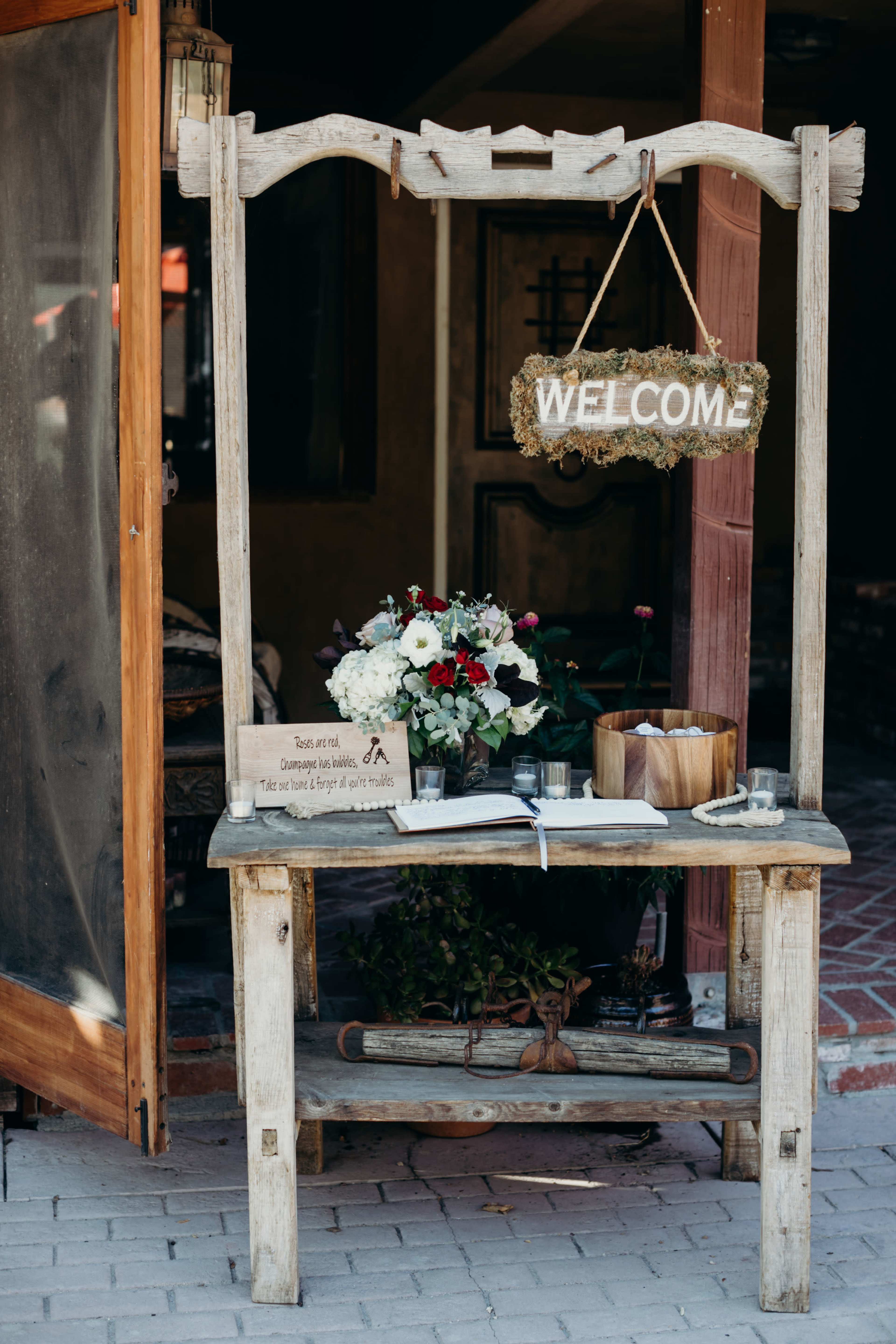 A rustic wooden welcome stand displays a bouquet of flowers and a sign in front of a doorway.