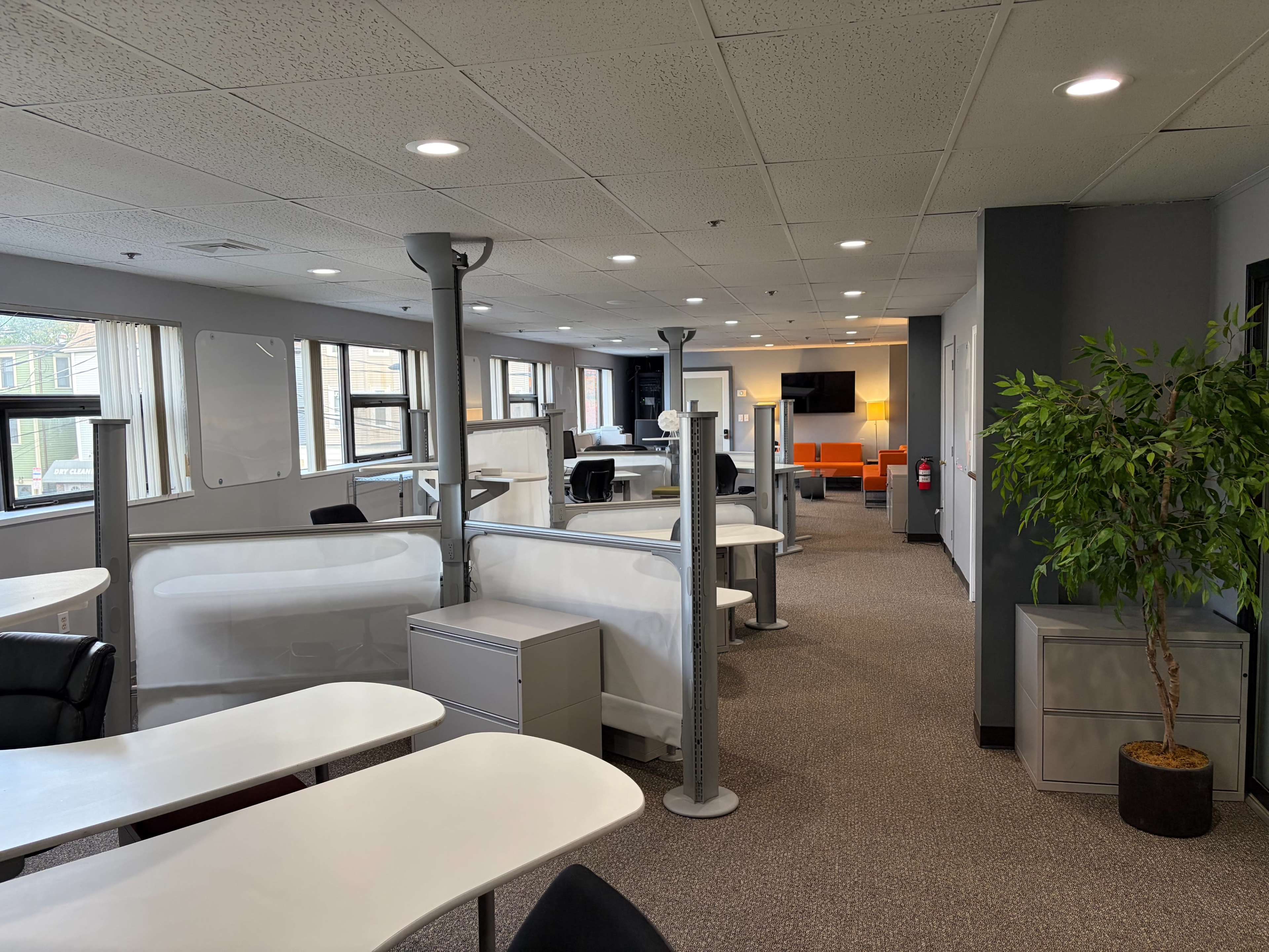 The image shows a modern office space with multiple workstations, a lounge area with orange seating, and large windows letting in natural light.