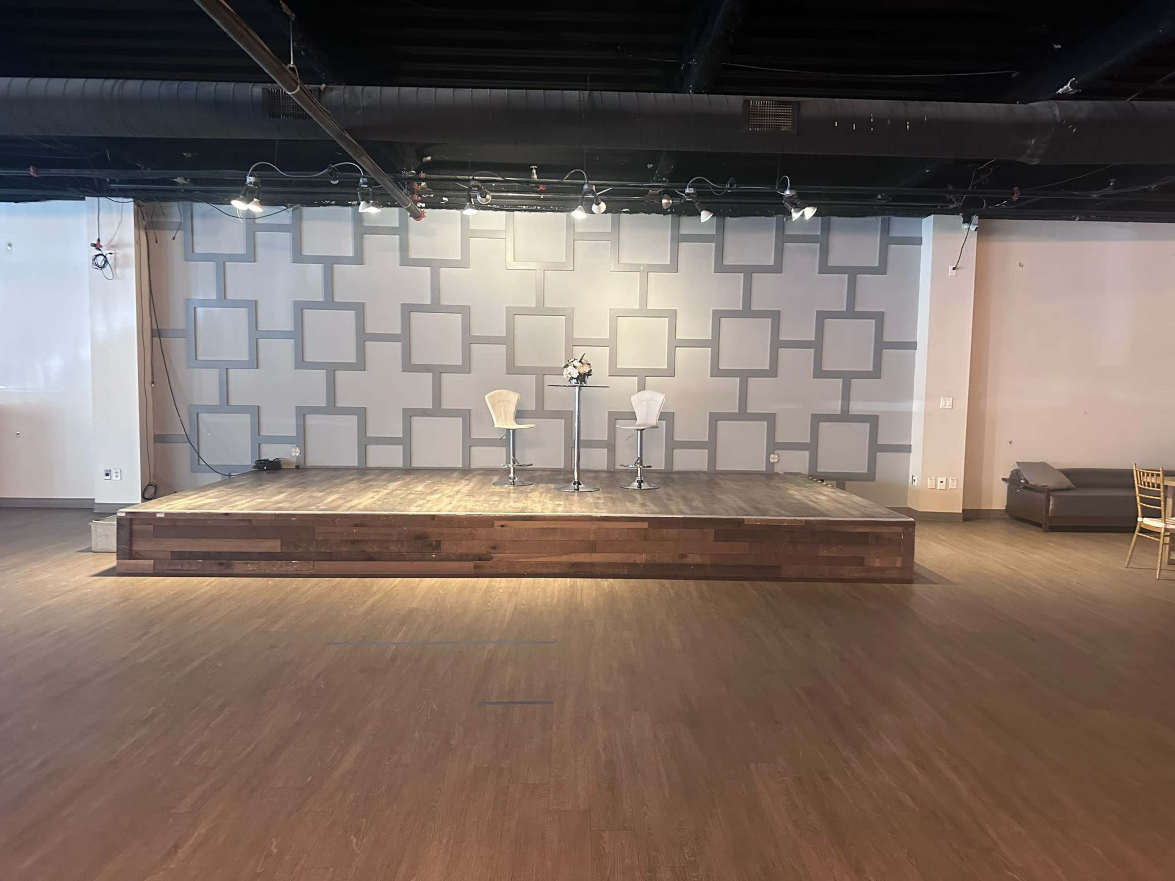 The image shows a wooden stage with two white chairs placed on it in a spacious room with a patterned wall and spotlights.