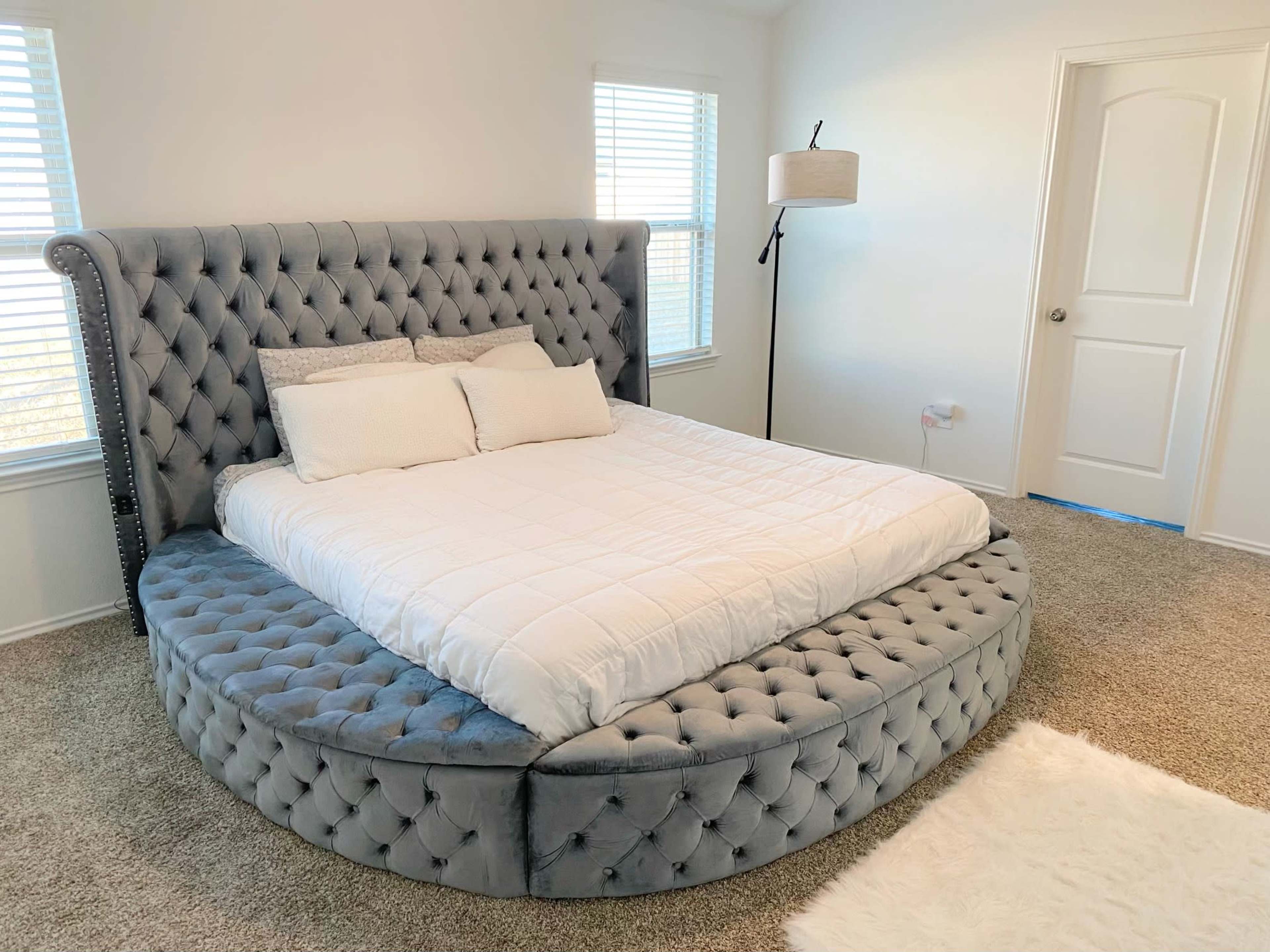 A round tufted bed with a gray fabric finish positioned in a well-lit bedroom with two windows and a floor lamp.