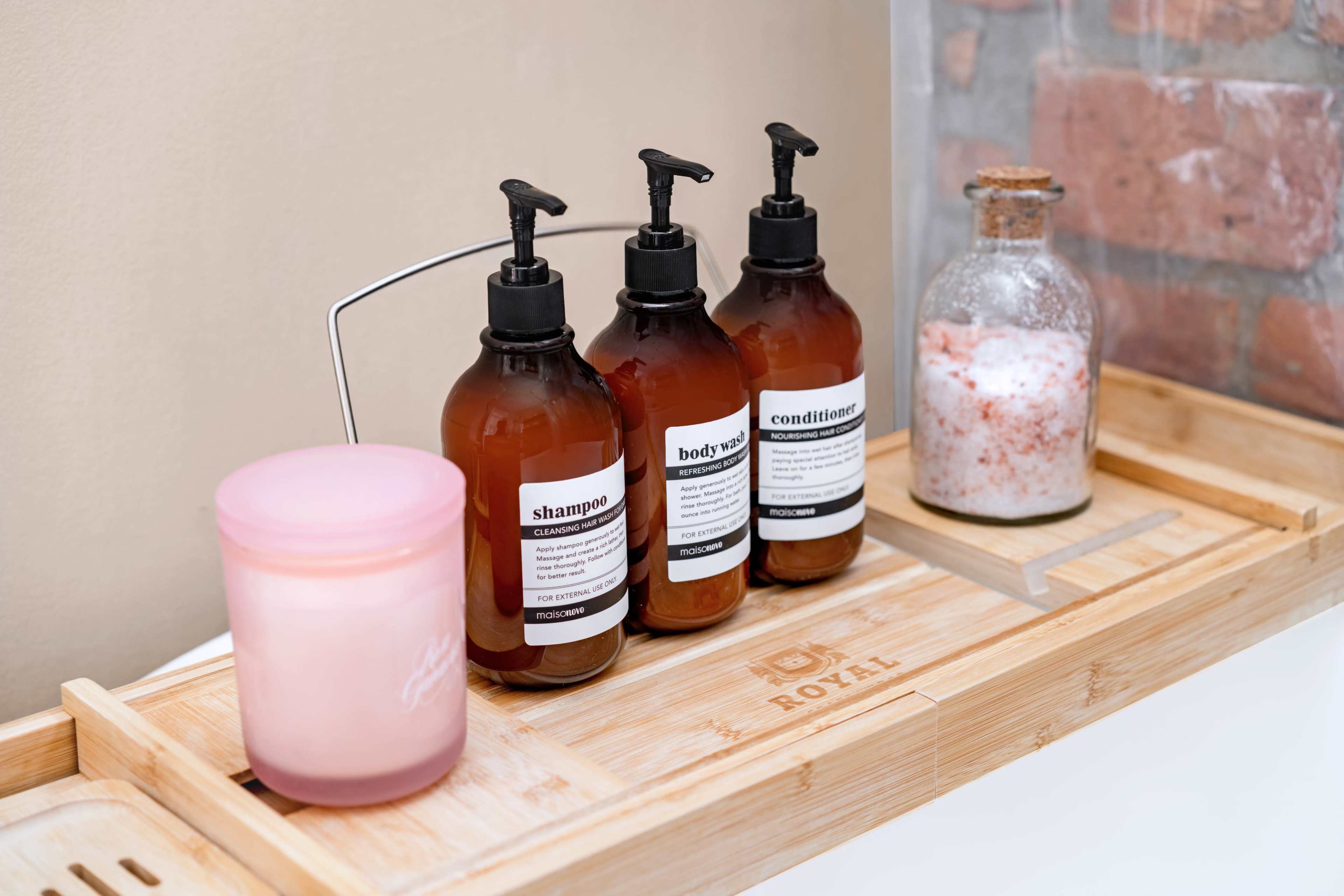 A bamboo tray holds three amber bottles labeled "shampoo," "conditioner," and "body wash," alongside a pink candle and a glass jar filled with salt.