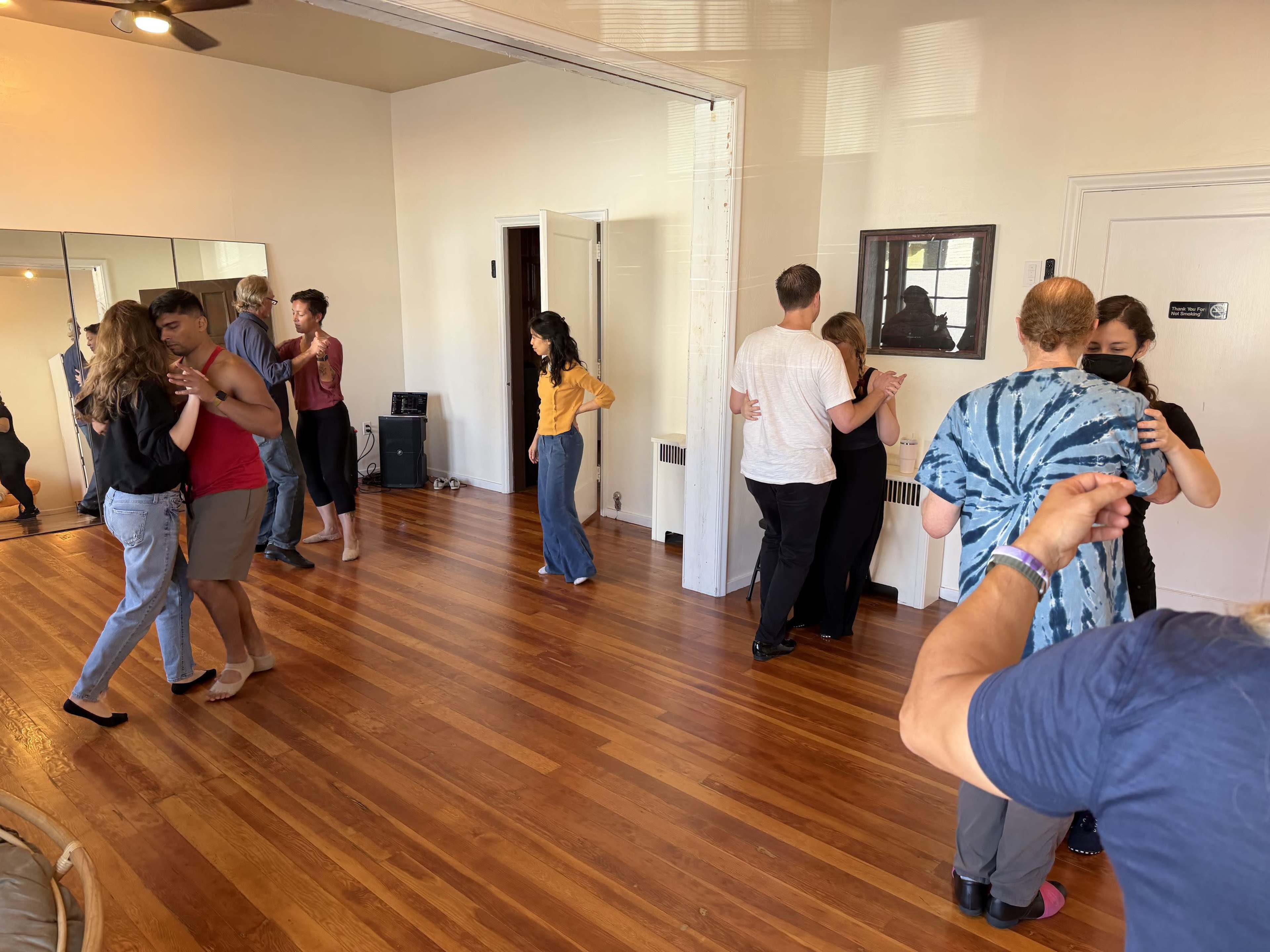 A group of people is dancing in a bright studio with wooden floors and mirrors on the walls.