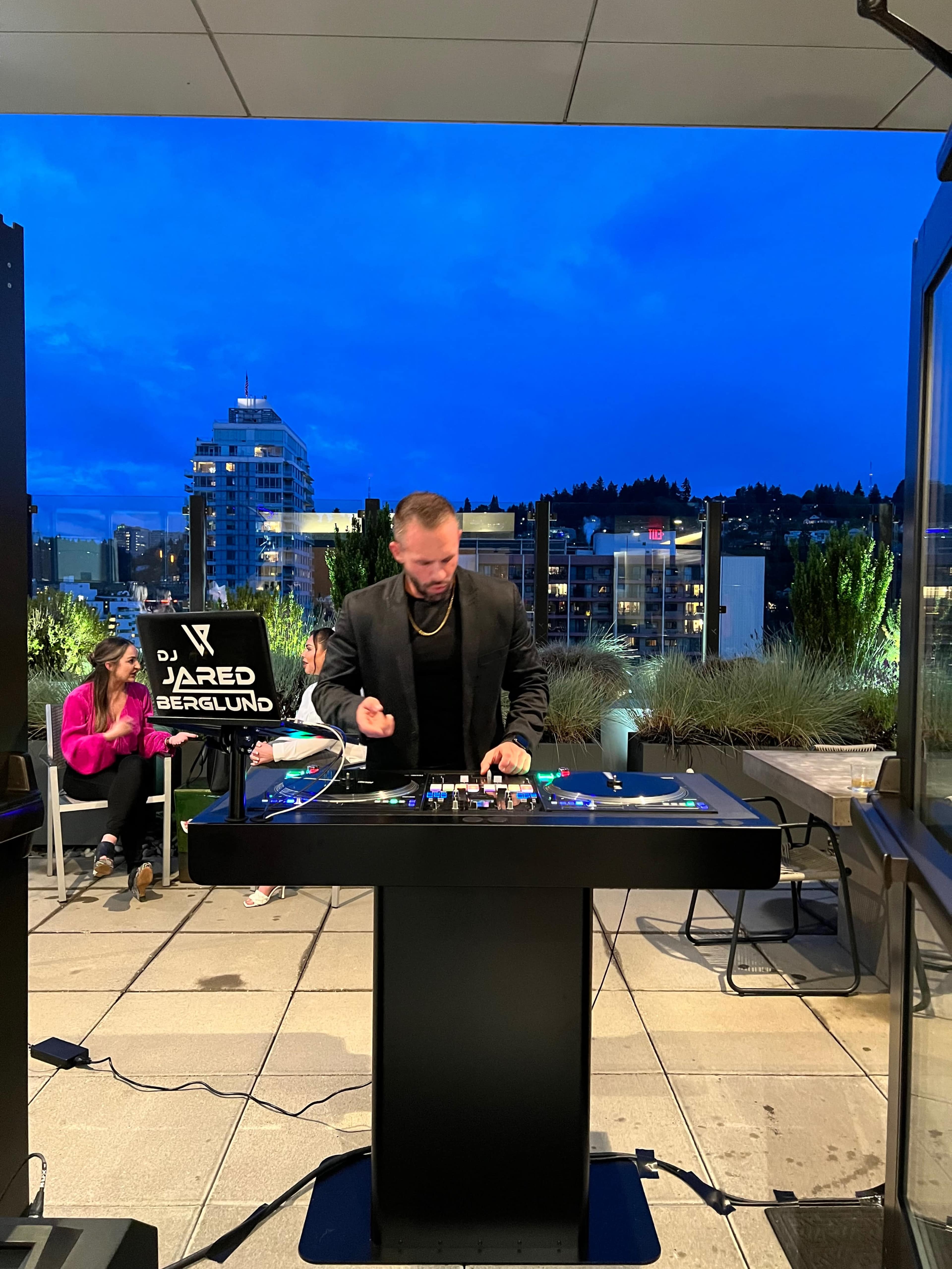 A DJ performs in a rooftop setting at dusk, with a city skyline and guests seated nearby.