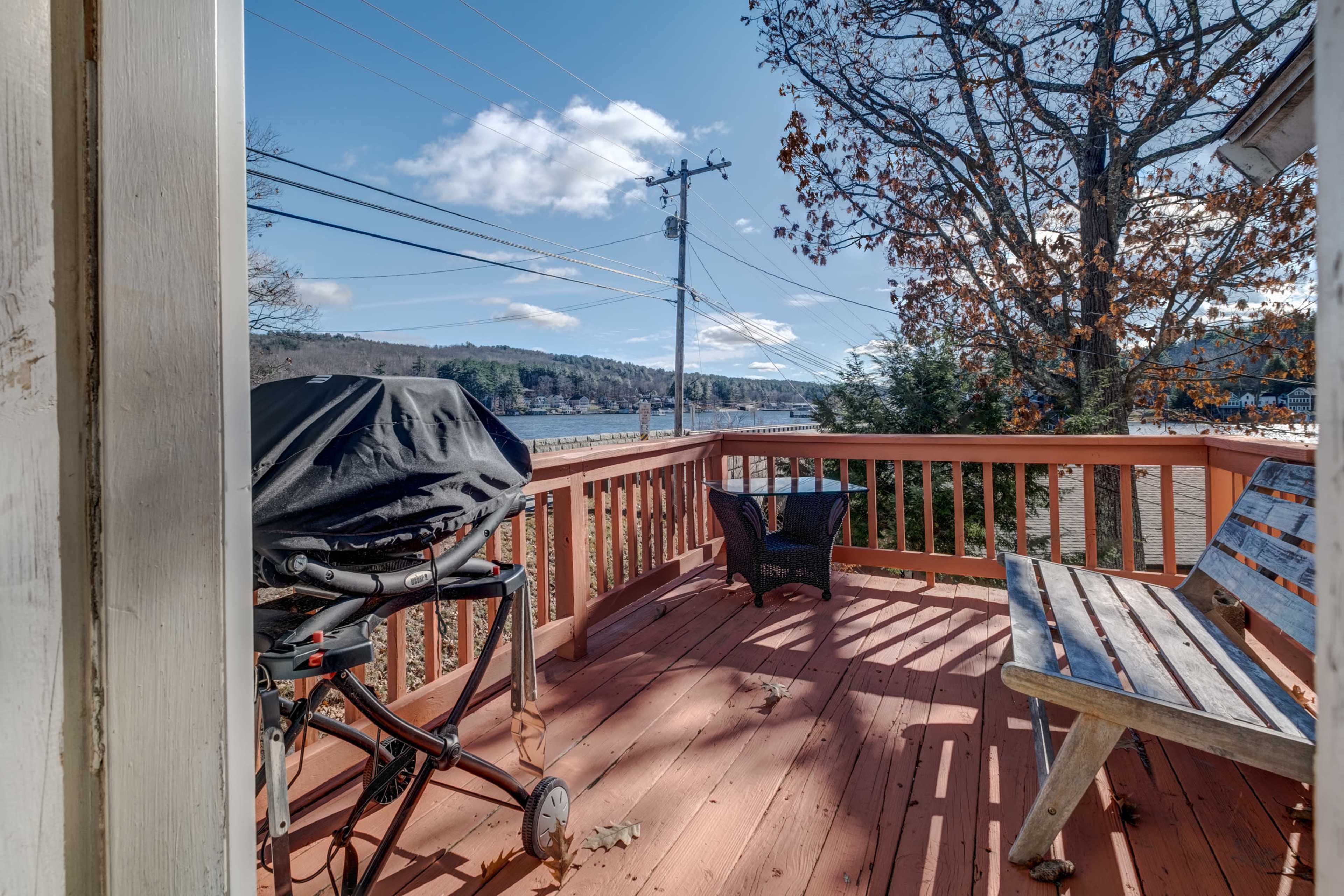 A wooden deck with a grill and a bench overlooks a lake and trees under a clear blue sky.