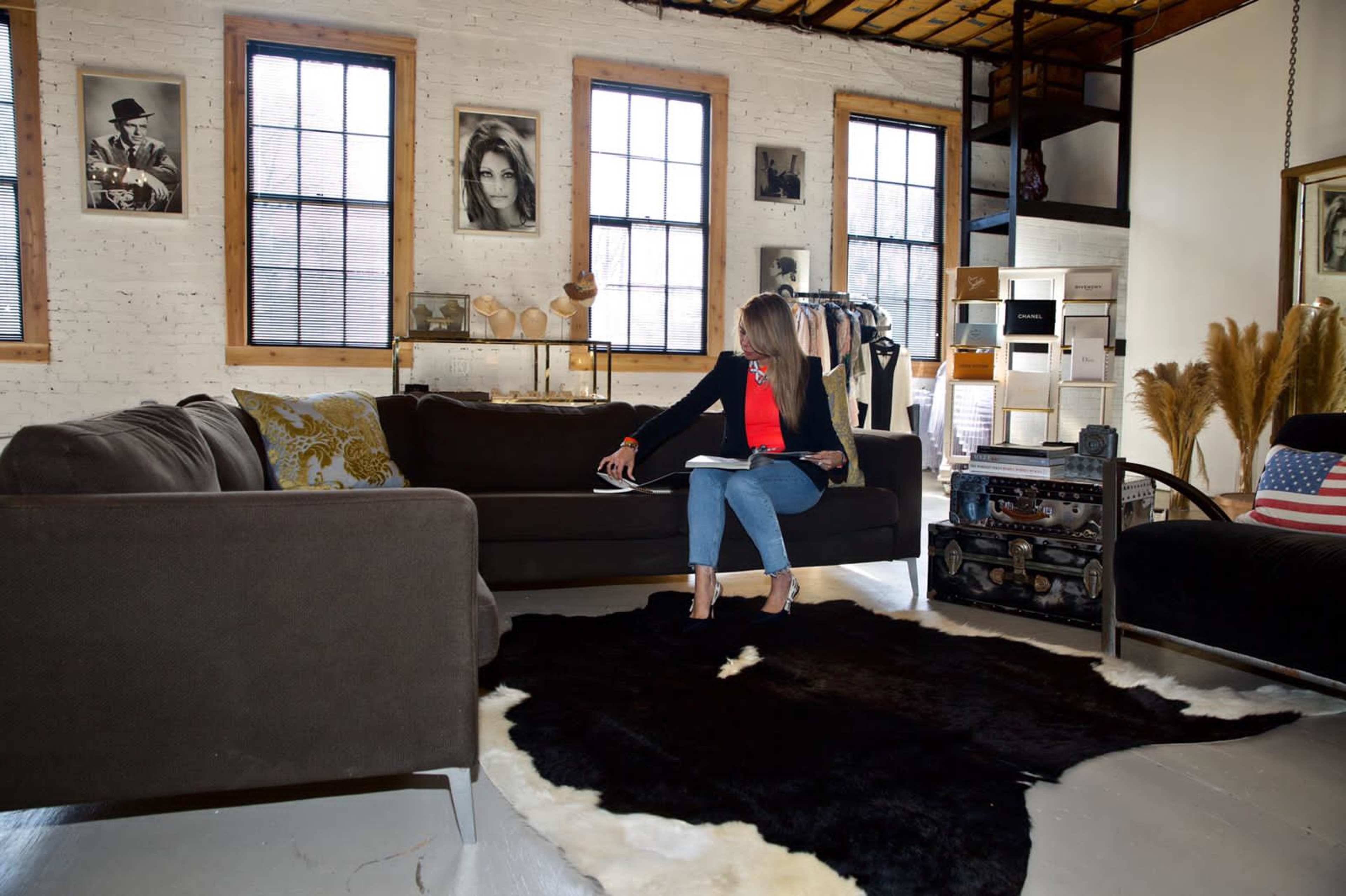 A woman is sitting on a dark sofa in a stylishly decorated room with large windows and various framed photographs on the walls.