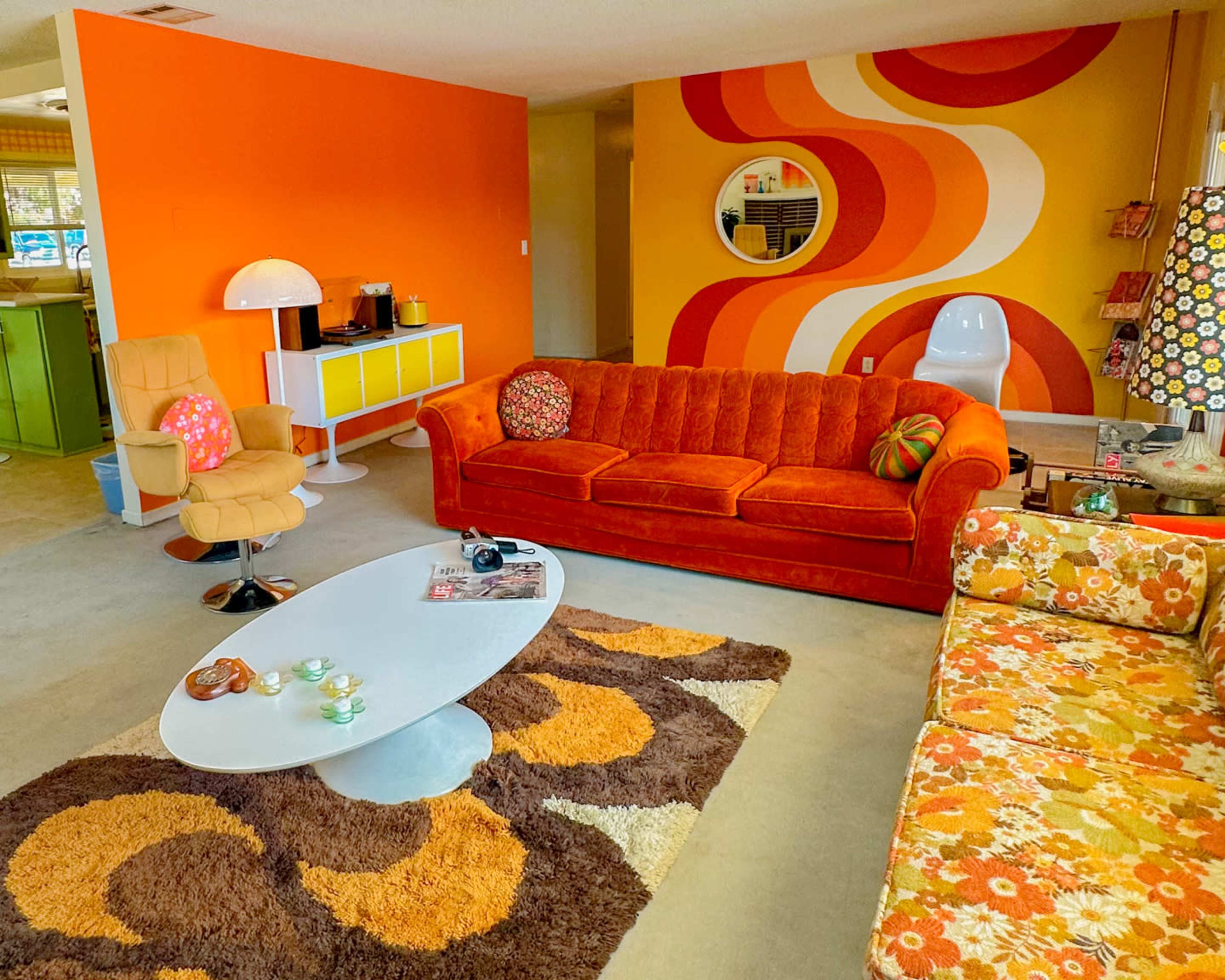 The living room features a vibrant orange and yellow color scheme with mid-century modern furniture, including a plush orange sofa and a patterned rug.