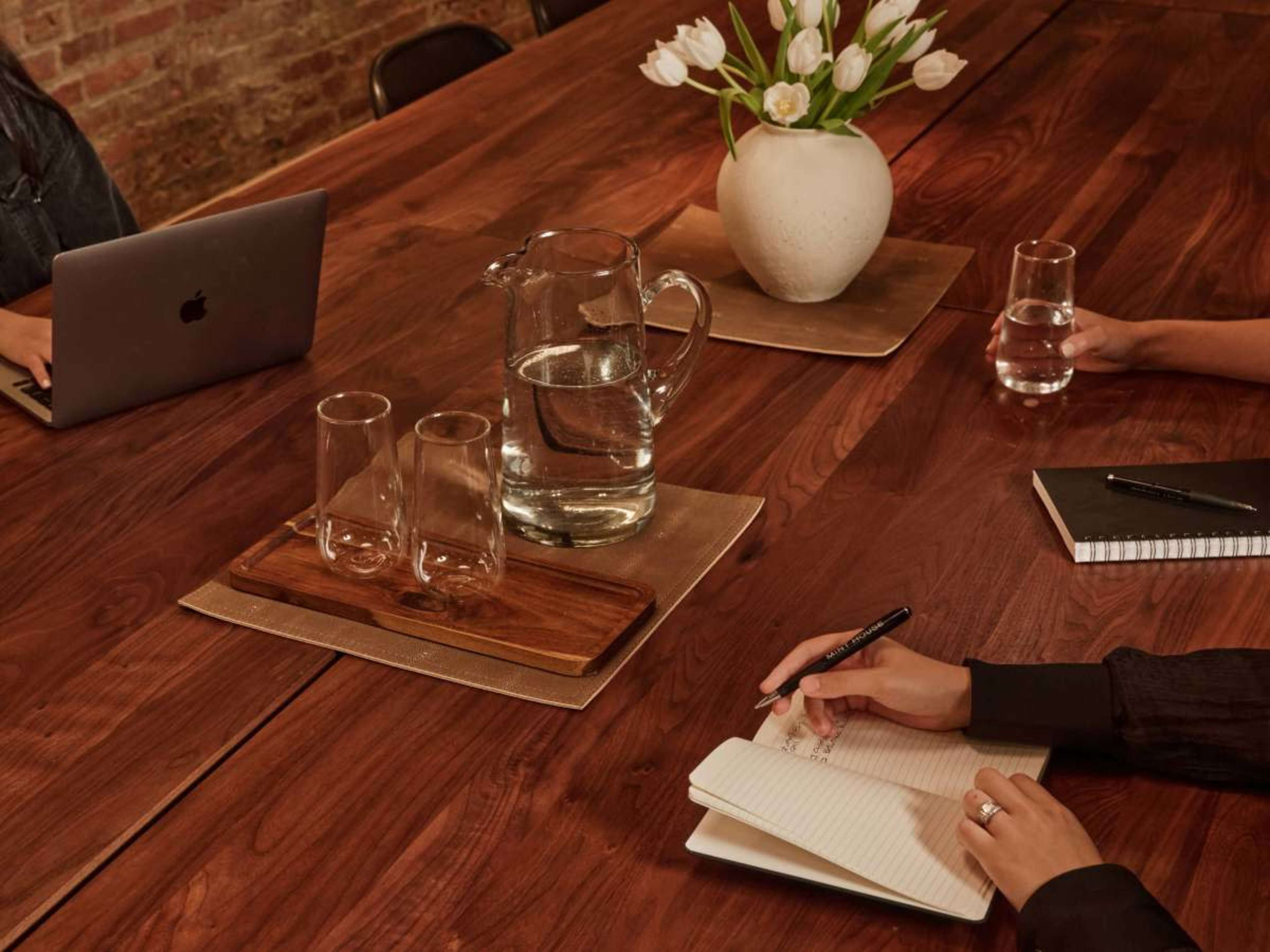 A wooden conference table is set with a water pitcher, two glasses, a notebook, and a pen, while individuals work on laptops and take notes.