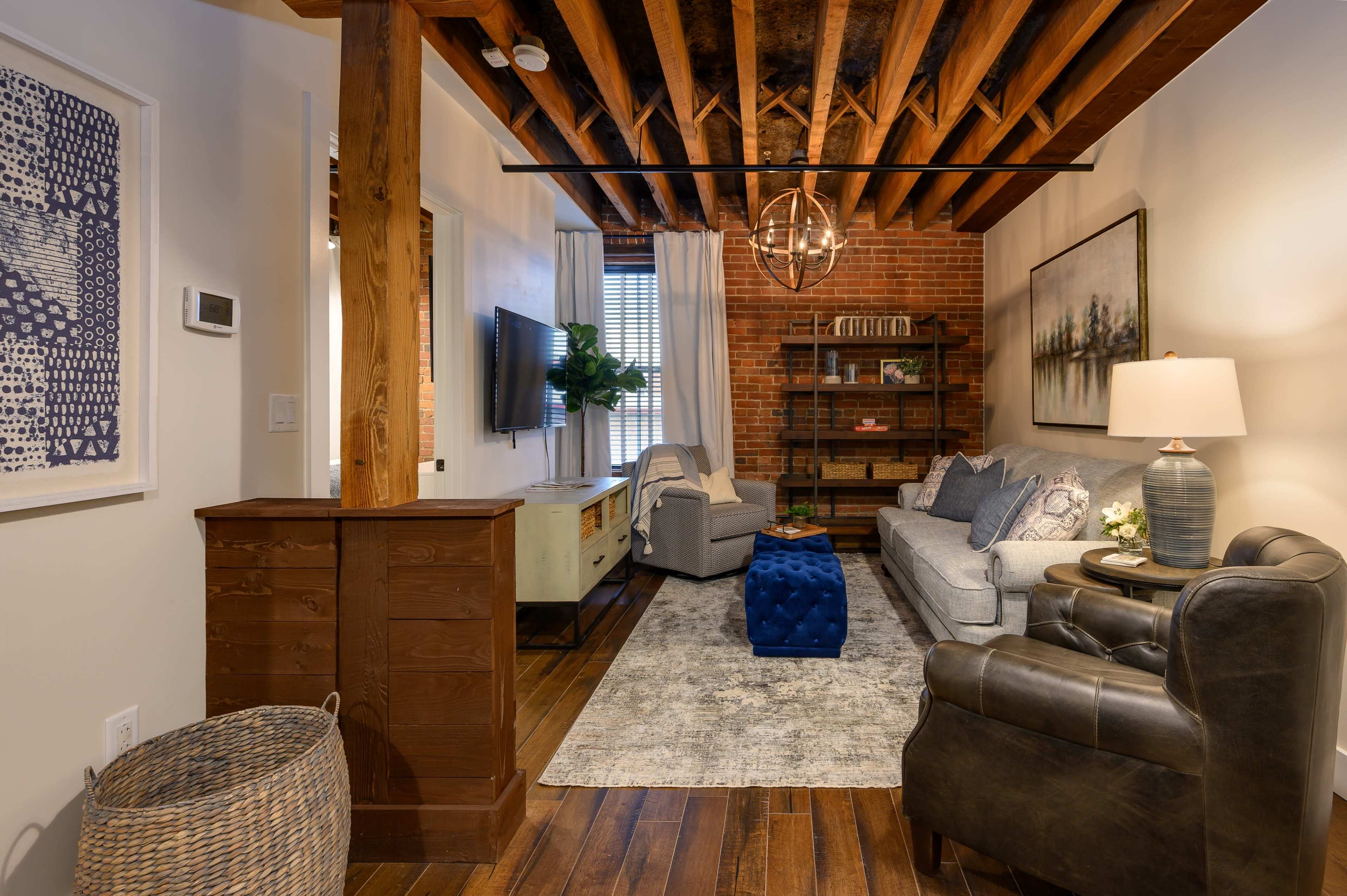 A living room with exposed wooden beams, a gray sofa, a blue ottoman, and a TV mounted on the wall, surrounded by brick and light-colored walls.