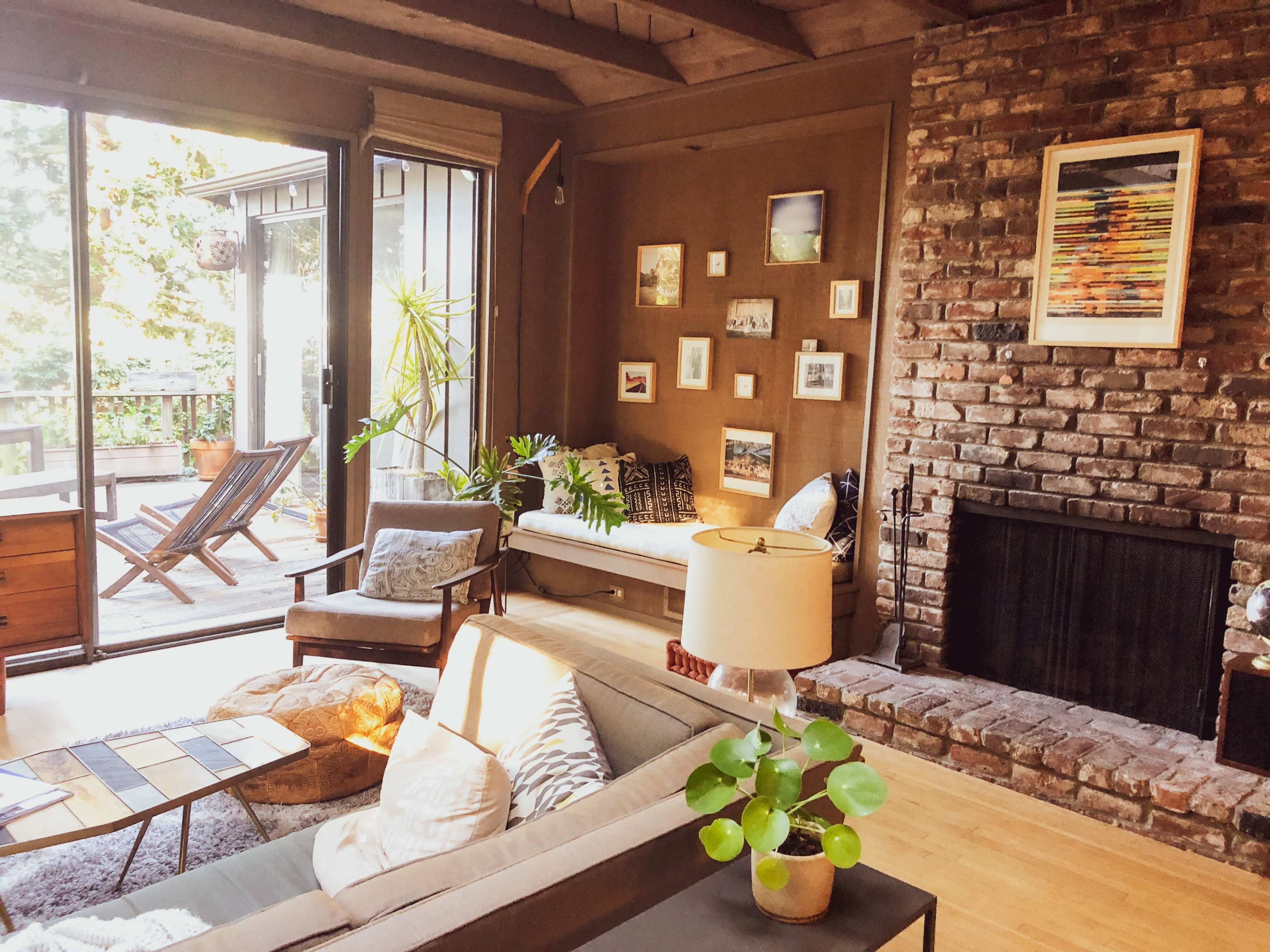 A cozy living room featuring a brick fireplace, a beige sofa with decorative pillows, and large windows leading to a deck.