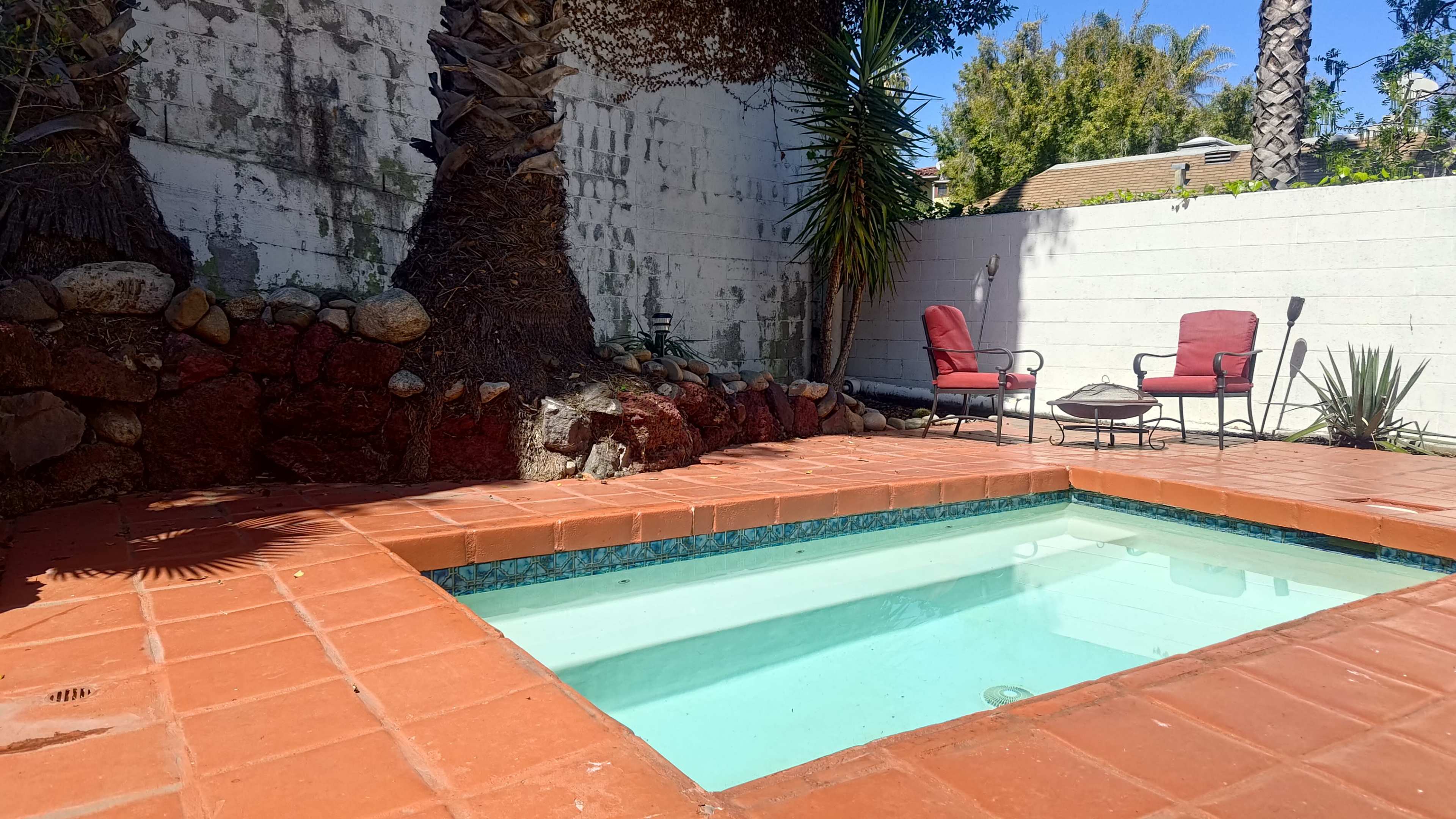 The image shows a small, empty swimming pool surrounded by a tiled patio, with two red chairs positioned nearby and palm trees in the background.