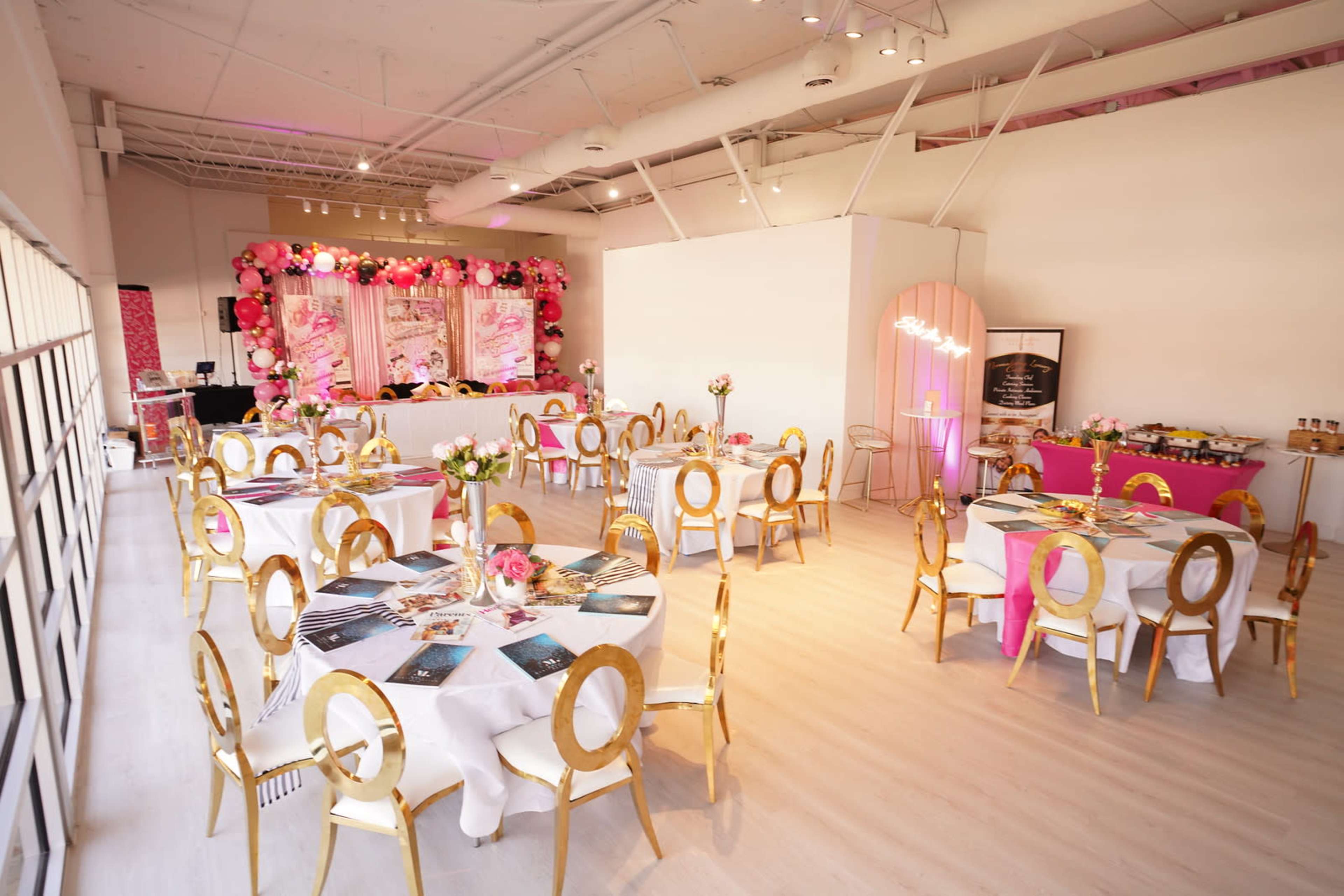 The image shows a well-decorated event space with round tables set for guests, featuring gold chairs and floral centerpieces, and a backdrop with pink decorations.