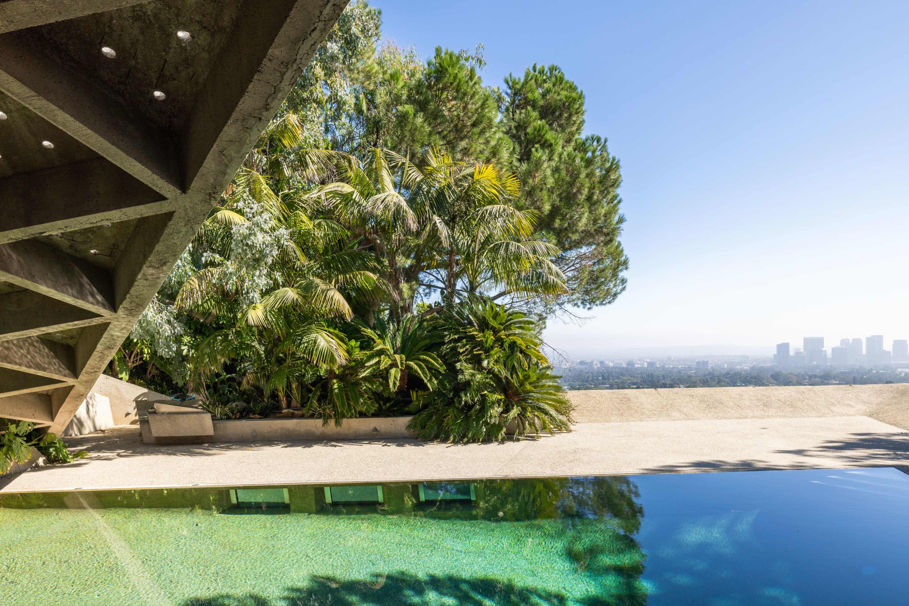 A modern architectural structure with an angled concrete overhang, overlooking a clear swimming pool and lush greenery that provides a view of the distant city skyline.