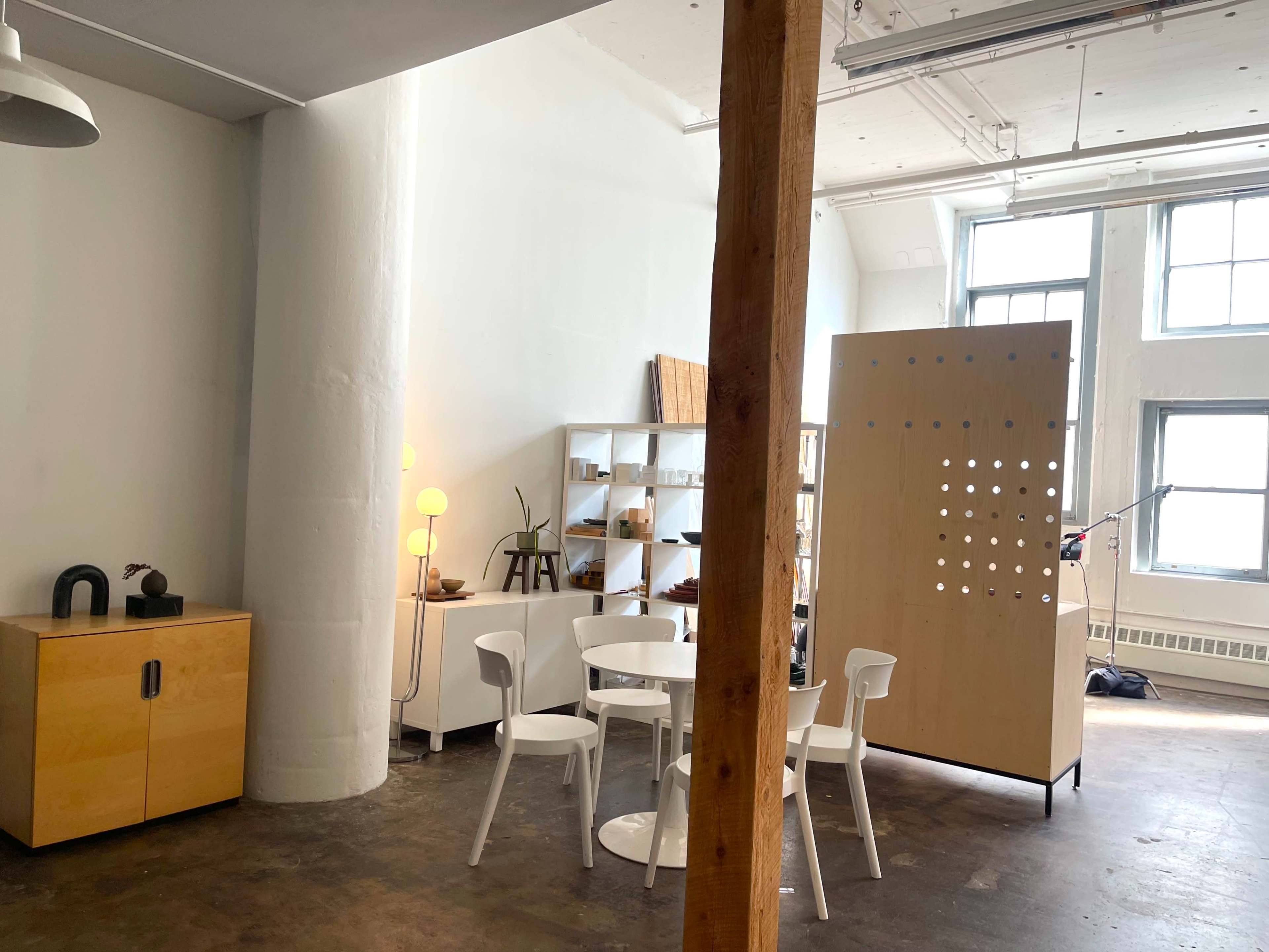 A modern, minimalist interior with a wooden support column, a circular dining table surrounded by white chairs, and a partitioned shelf displaying various items.