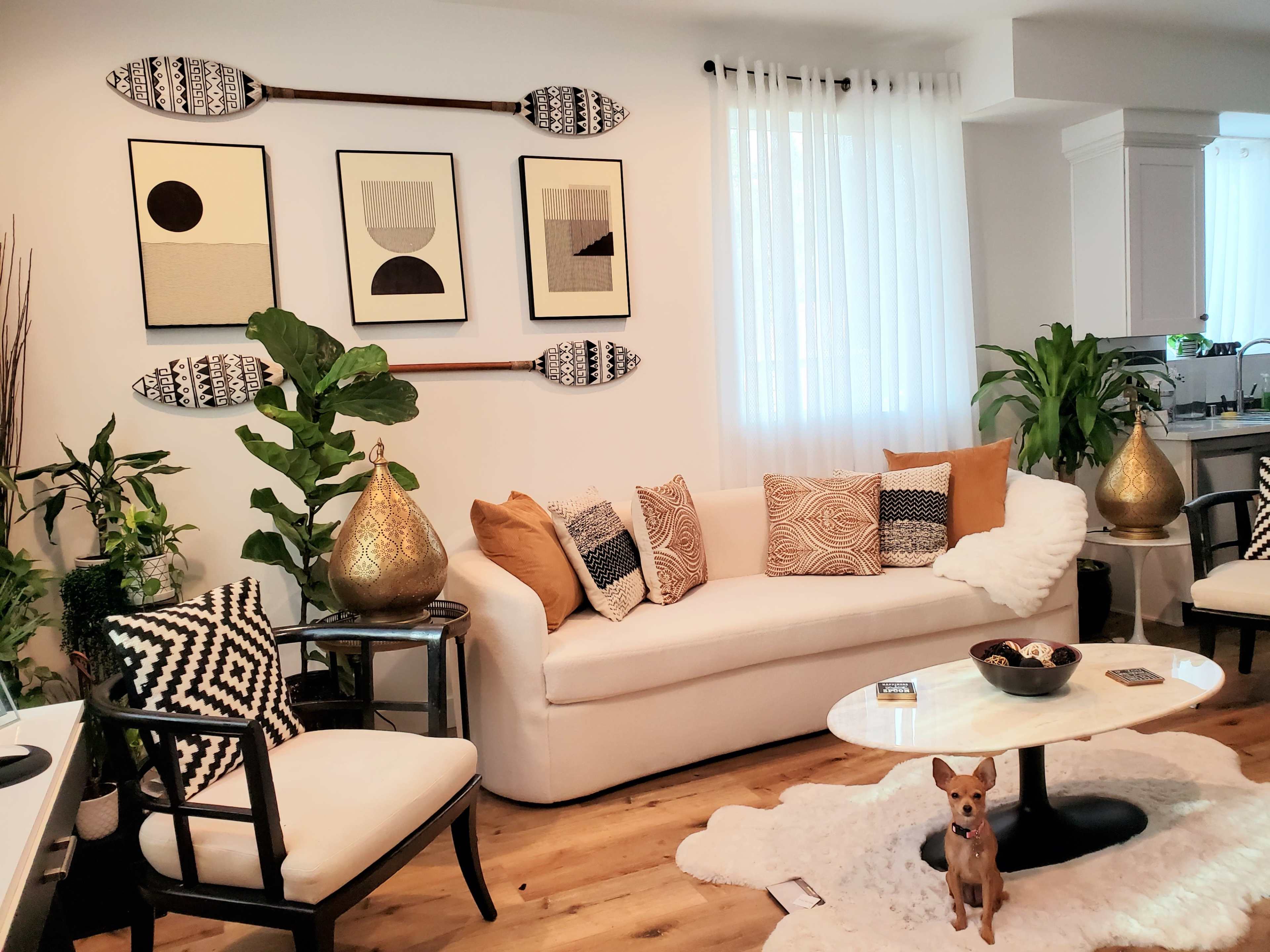 The living room features a white sofa adorned with decorative pillows, paired with a black and white chair, surrounded by plants and artwork on the walls.