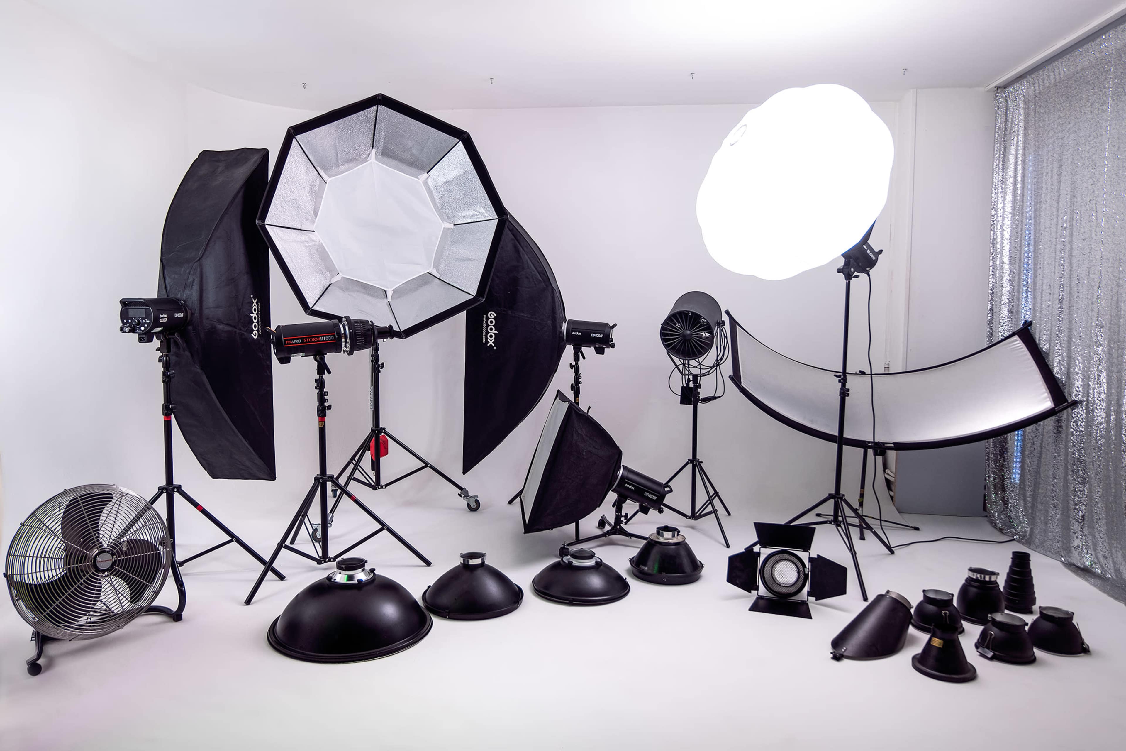 The image shows a photography studio setup featuring various lighting equipment, softboxes, and reflectors arranged on a white backdrop.