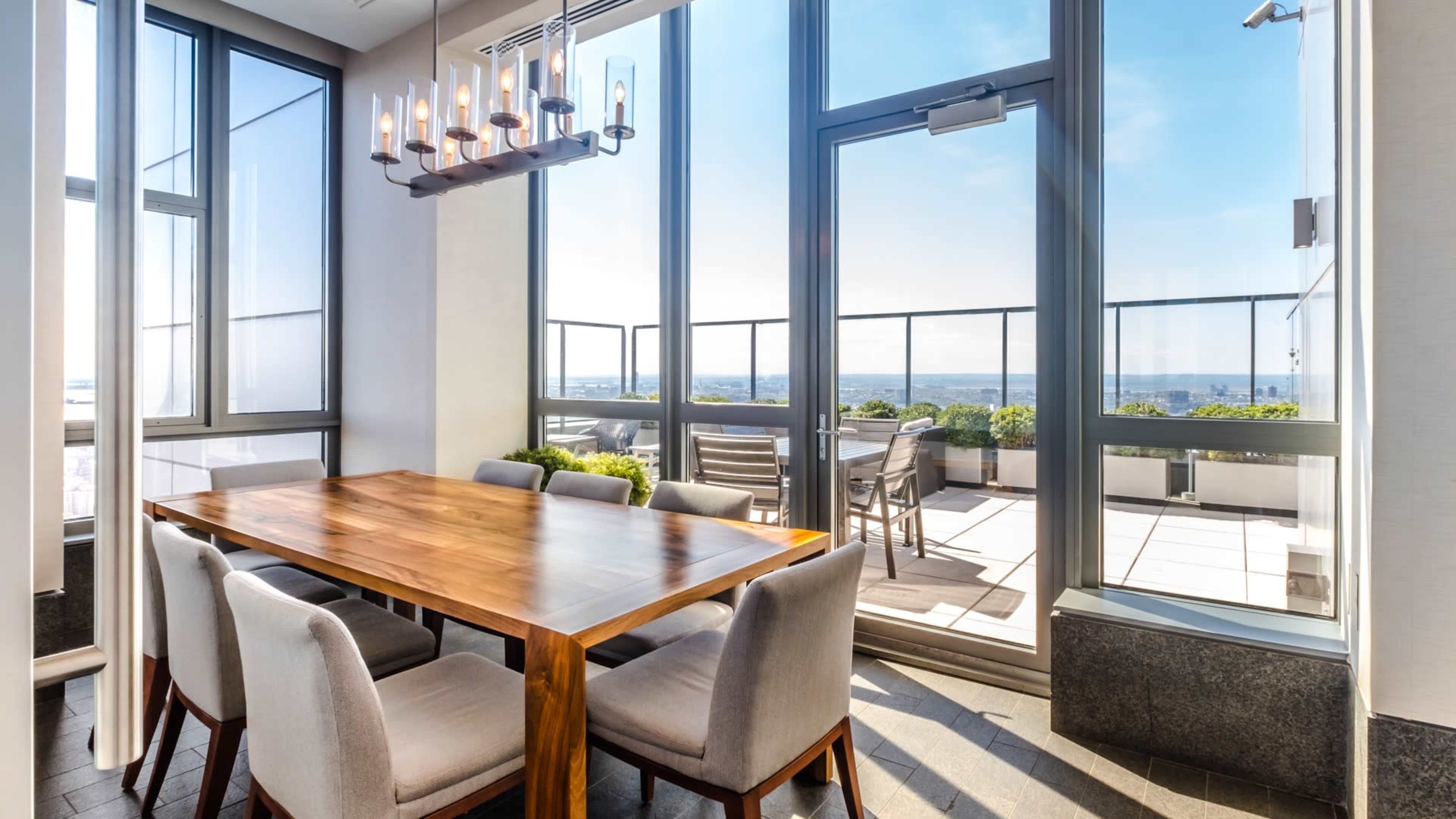 A dining area features a wooden table surrounded by upholstered chairs, with large windows offering a view of an outdoor terrace.
