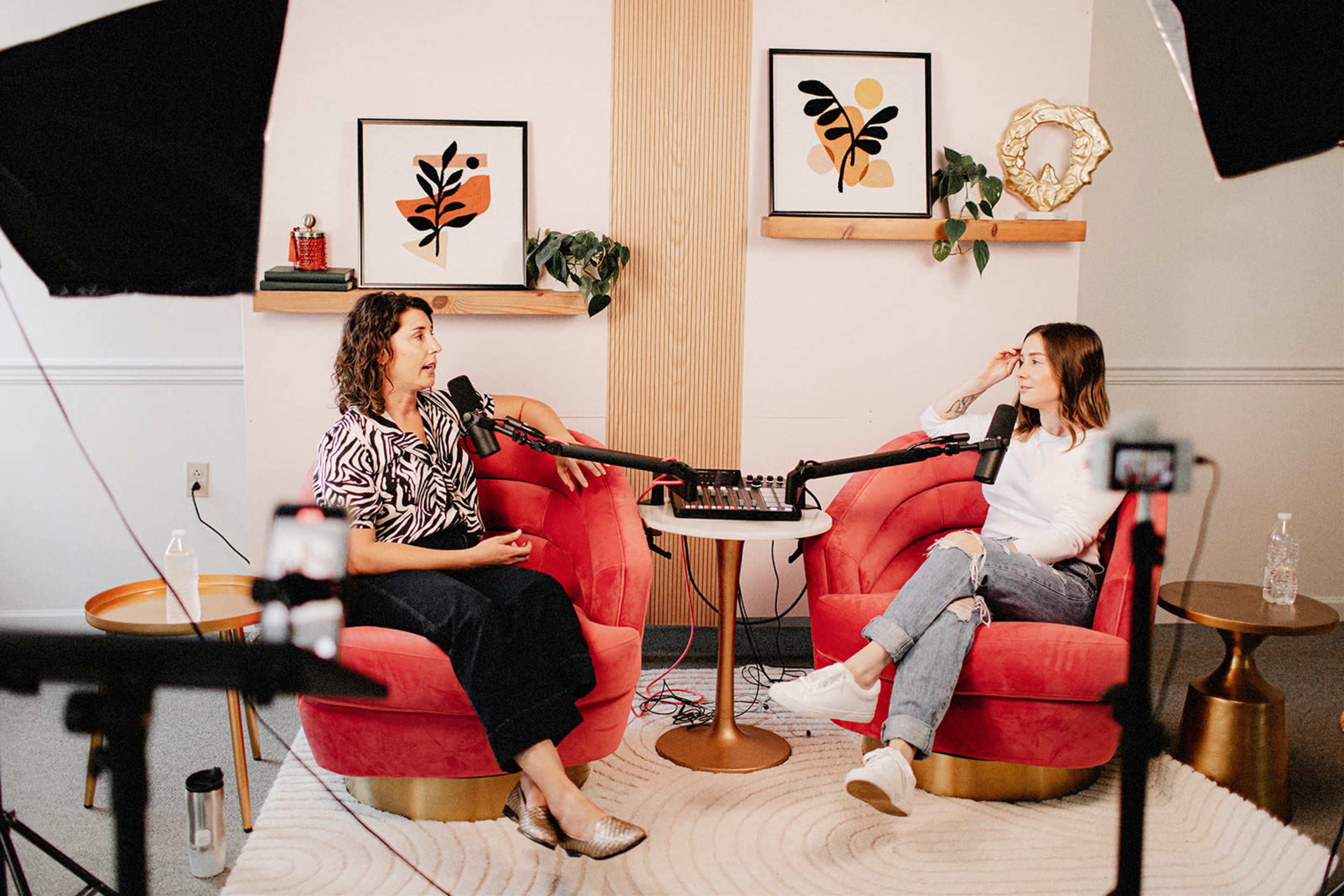 Two women are seated in a cozy studio environment with microphones in front of them, engaged in conversation.